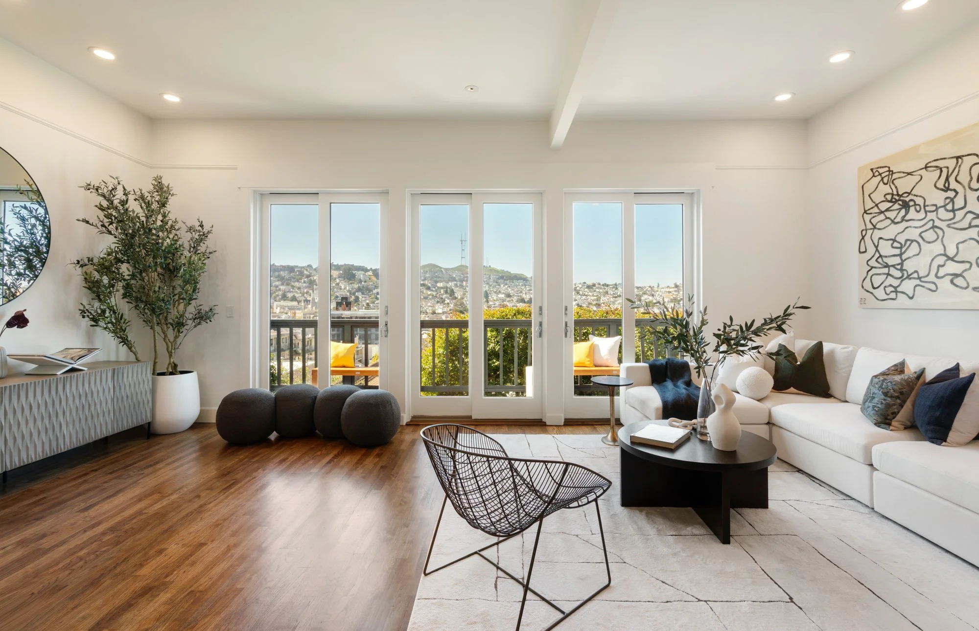 Bright living room with large glass doors leading to a balcony, wooden floors, a white sofa with pillows, a black coffee table with decorative vases, a wire chair, a large potted plant, and abstract wall art.