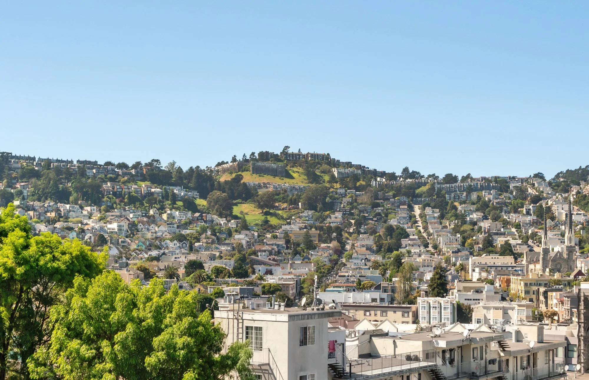 Cityscape of hills with residential buildings, lush green trees, and a bright blue sky.