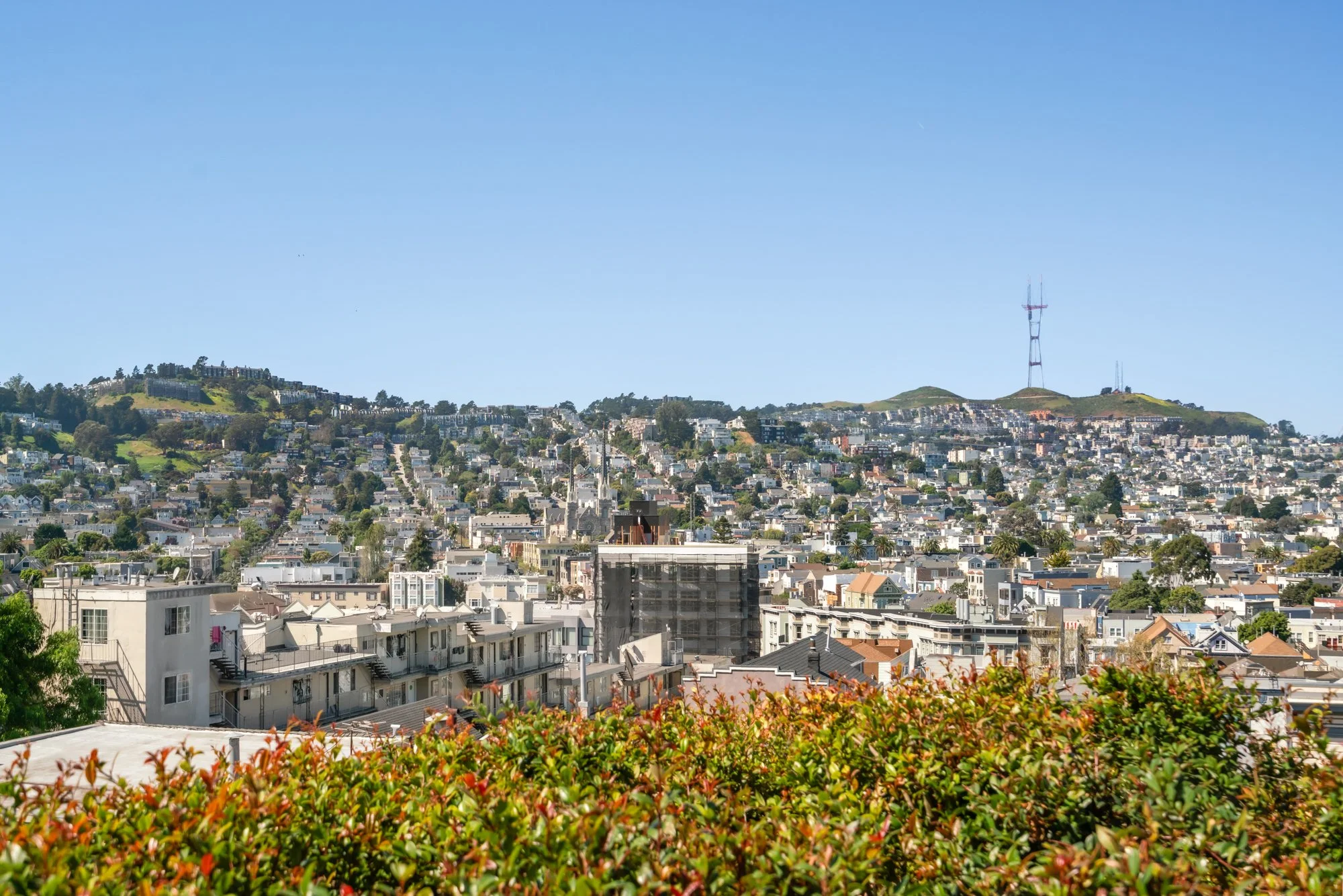 A cityscape of San Francisco with many residential buildings, hills, and a tall radio tower in the background under a clear blue sky.
