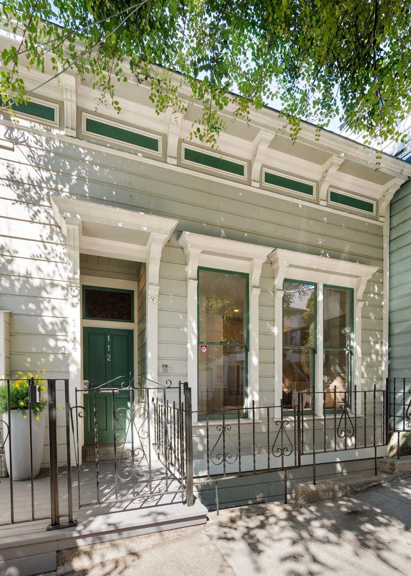 Front view of a Victorian-style house with light gray siding, green accents on the door and windows, decorative trim, and a small fenced porch. Shadows of tree leaves are cast on the house.