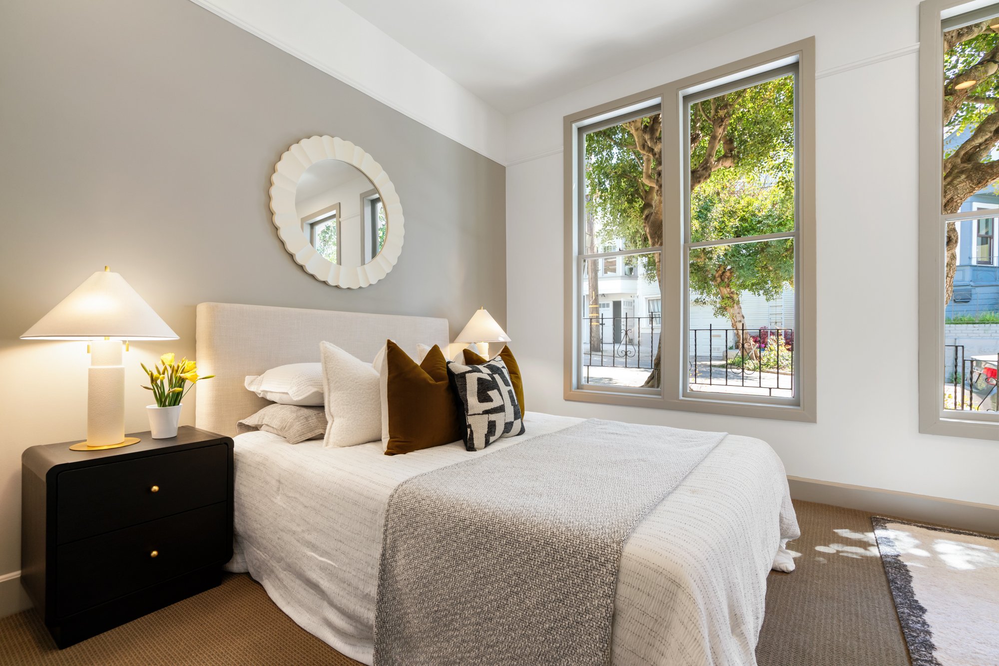 Bright bedroom with large windows showing trees outside, beige carpet, white bed with pillows, bedside table with lamp and potted plant, mirror on wall.