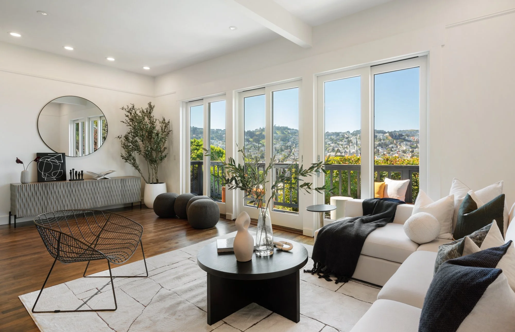 Bright living room with white walls, large windows overlooking a cityscape, a white sofa with black, white, and gray pillows, a black coffee table, decorative vases, a patterned sideboard, and indoor plants.