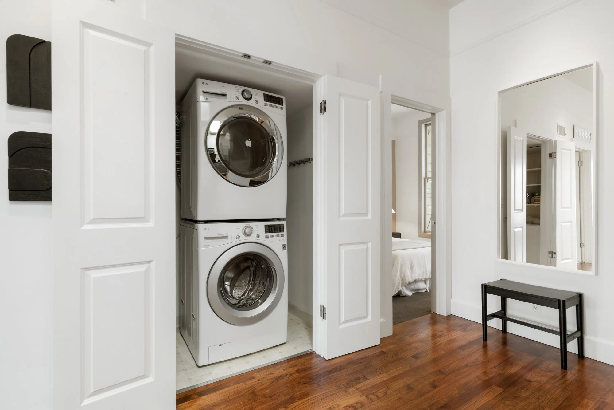 Stacked front-loading washing machines in laundry closet with white doors open, hardwood floor, and adjacent bedroom with bed and window.