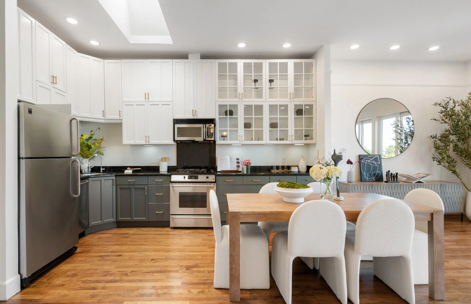 Modern kitchen with white upper cabinets, gray lower cabinets, black countertops, stainless steel refrigerator, microwave, and stove. A wooden dining table with six white upholstered chairs, decorative vases, and flowers. Wooden flooring, round mirro
