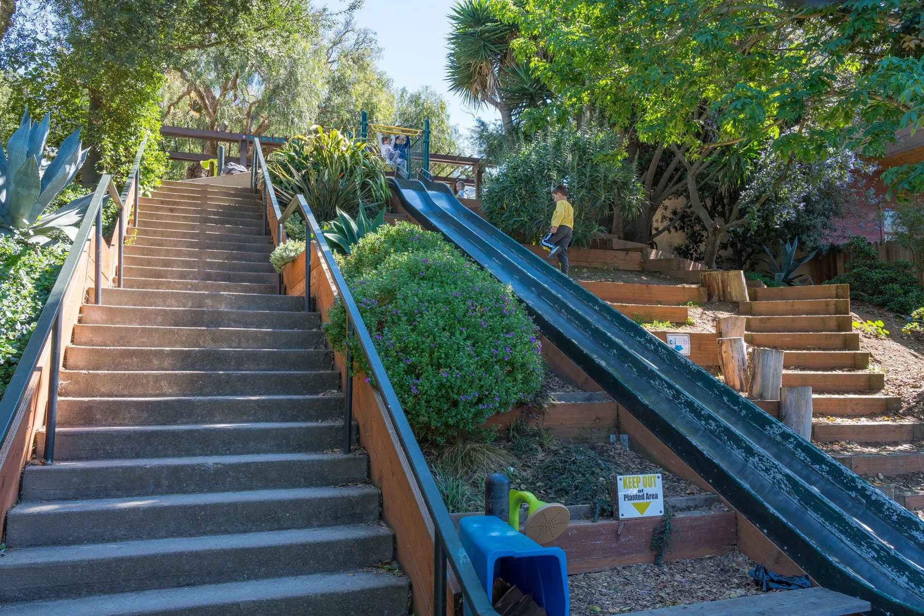 A staircase and a slide in a backyard with lush greenery and trees. Children are playing near the slide, and there is a sign that says 'Keep Out Planted Area' at the bottom.