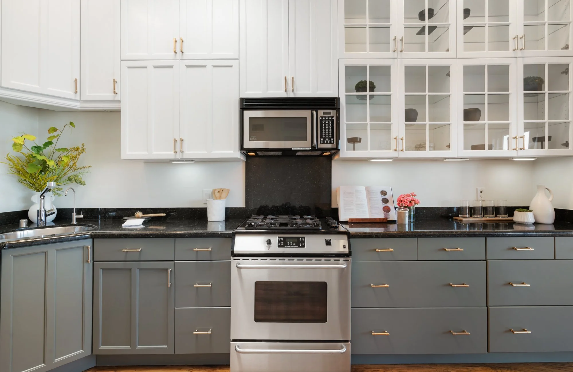 Modern kitchen with white cabinets, black countertops, and grey lower cabinets, with a stainless steel stove and microwave, and decorative items like flowers and pitchers.