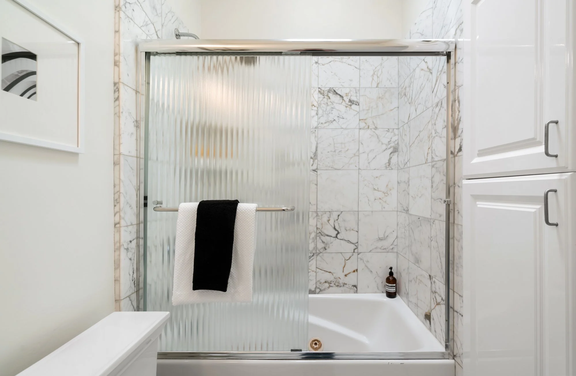 Bathroom shower with marble tiles, a sliding glass door with frosted glass, a towel rack with black and white towels, a bottle of soap or shampoo on the shower floor, white cabinetry, and a white wall with framed artwork.