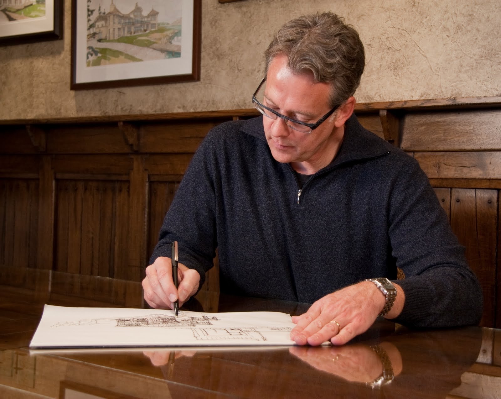 Wayne Visbeen is sitting at a wooden table, sketching architectural designs on a sketch pad with a pen. The background includes framed pictures on a beige and wooden-paneled wall.
