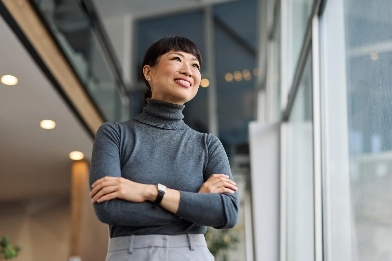 A woman with short dark hair in a gray turtleneck sweater standing near a large window, smiling and looking outside.
