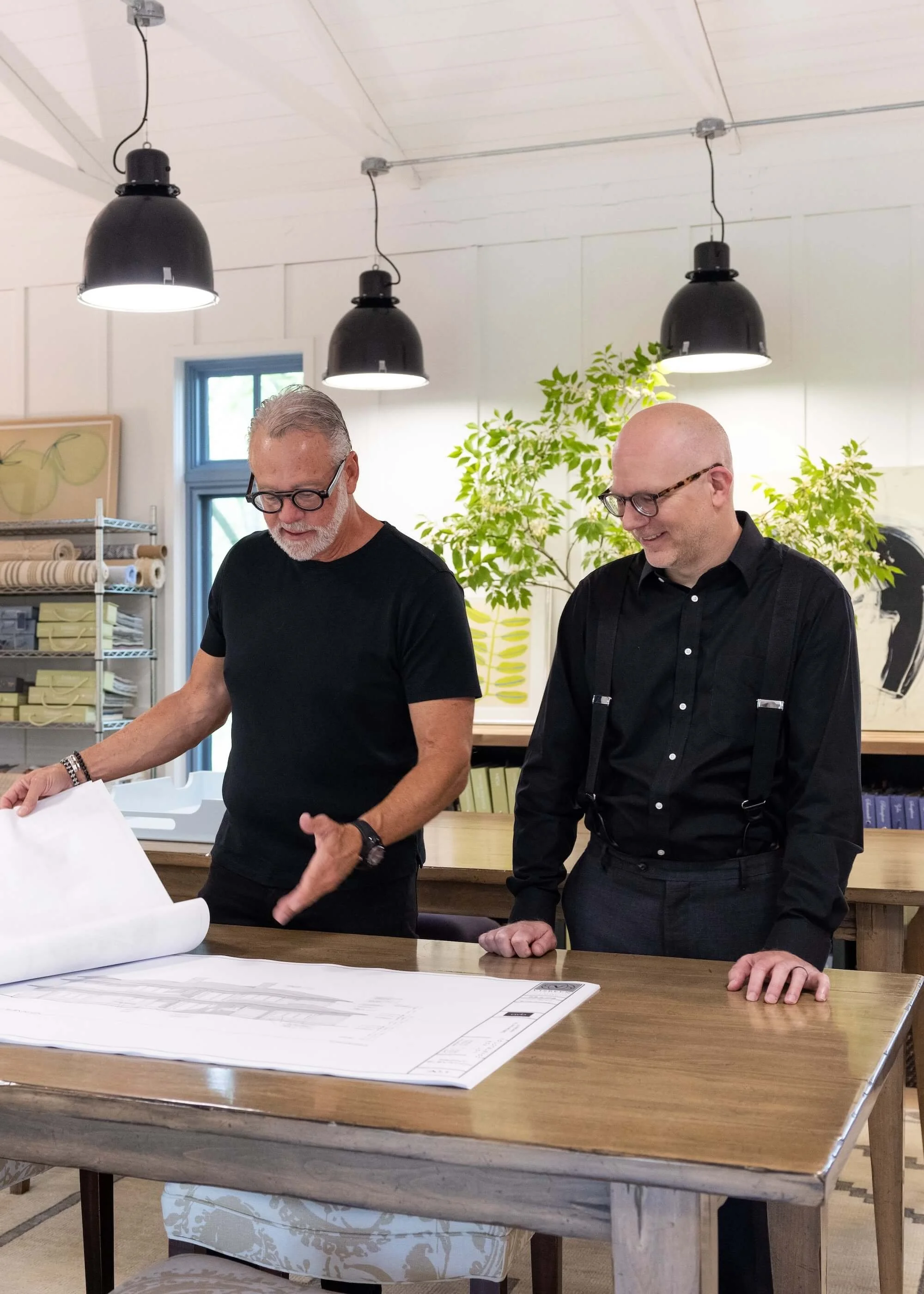 Two men in black shirts and glasses standing at a large table with architectural or engineering plans, in a well-lit room with black pendant lights, plants, and artwork on the walls.