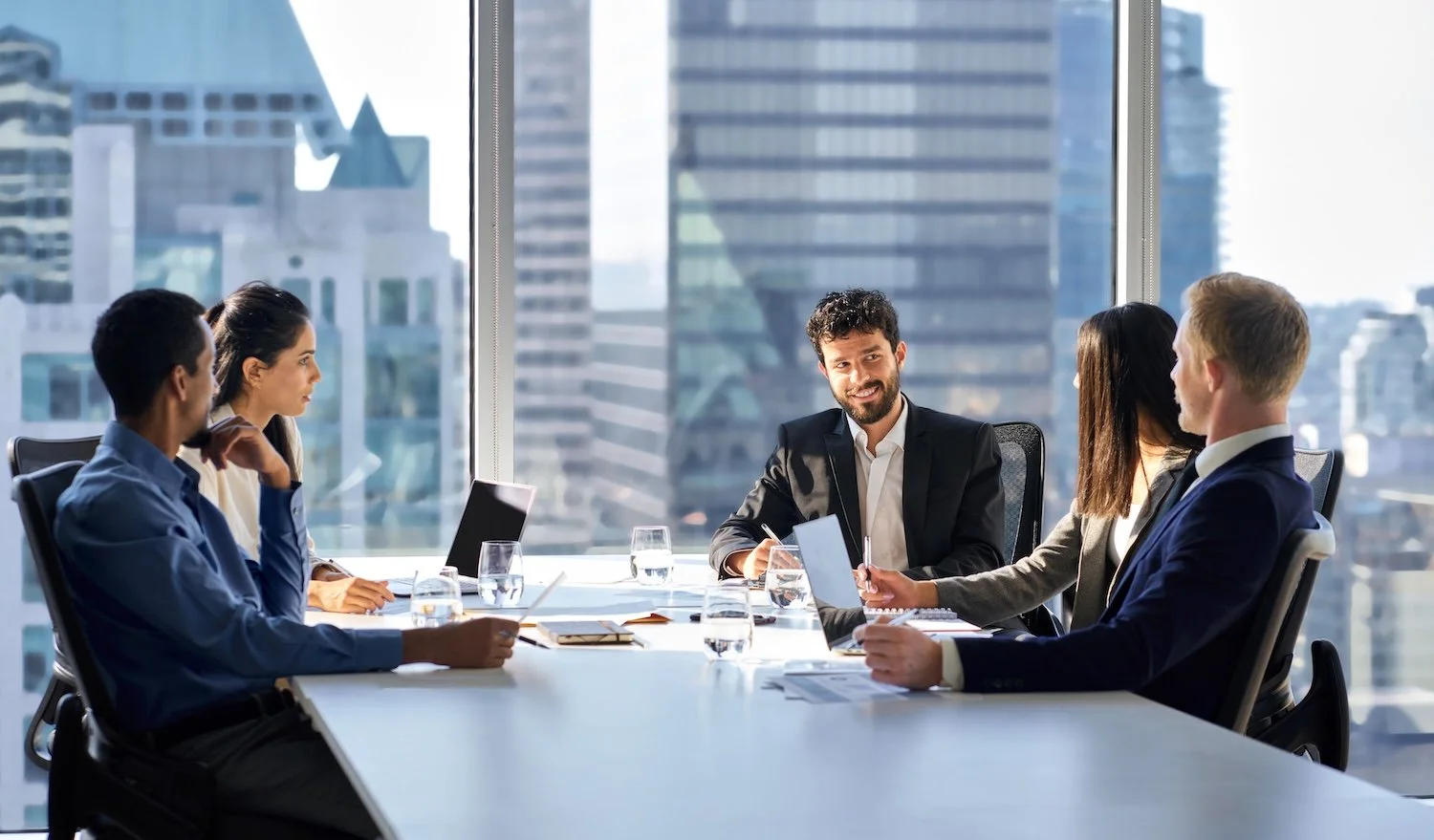 Business meeting with five people in a modern office with large windows and city view.