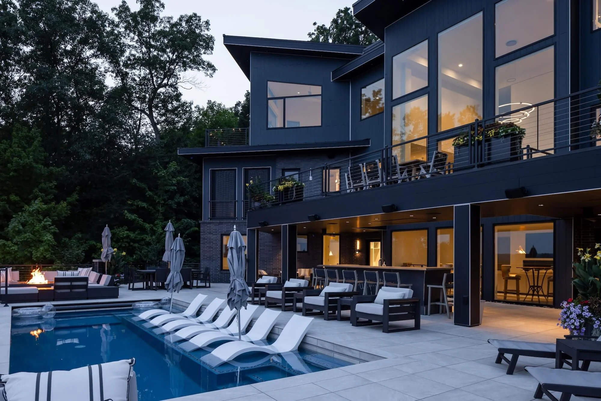Modern house with large glass windows, outdoor patio with seating, pool, and firepit, surrounded by trees at dusk.