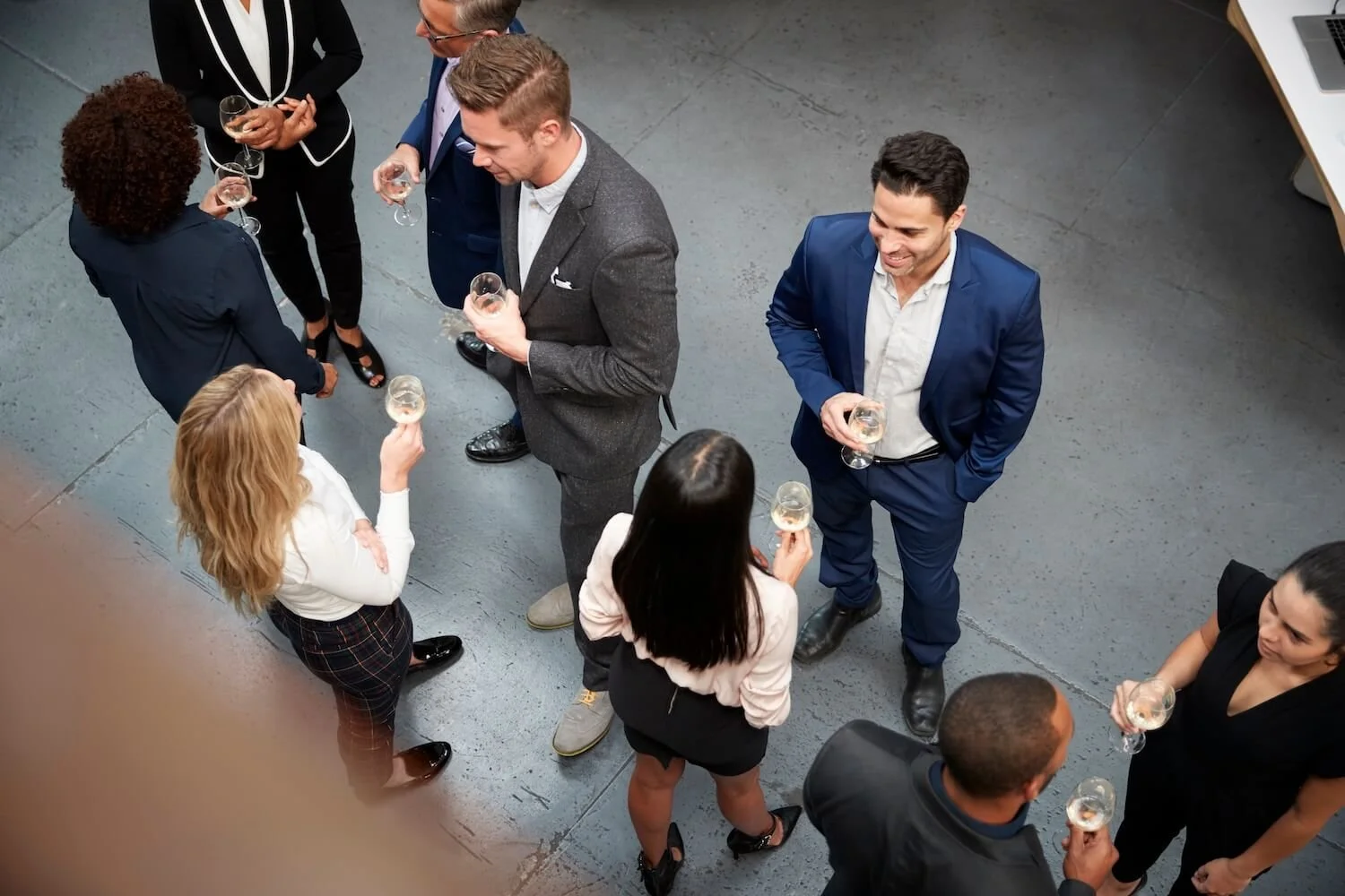 Group of seven people at a social gathering, some holding glasses of champagne, standing and talking in an indoor space with gray flooring.
