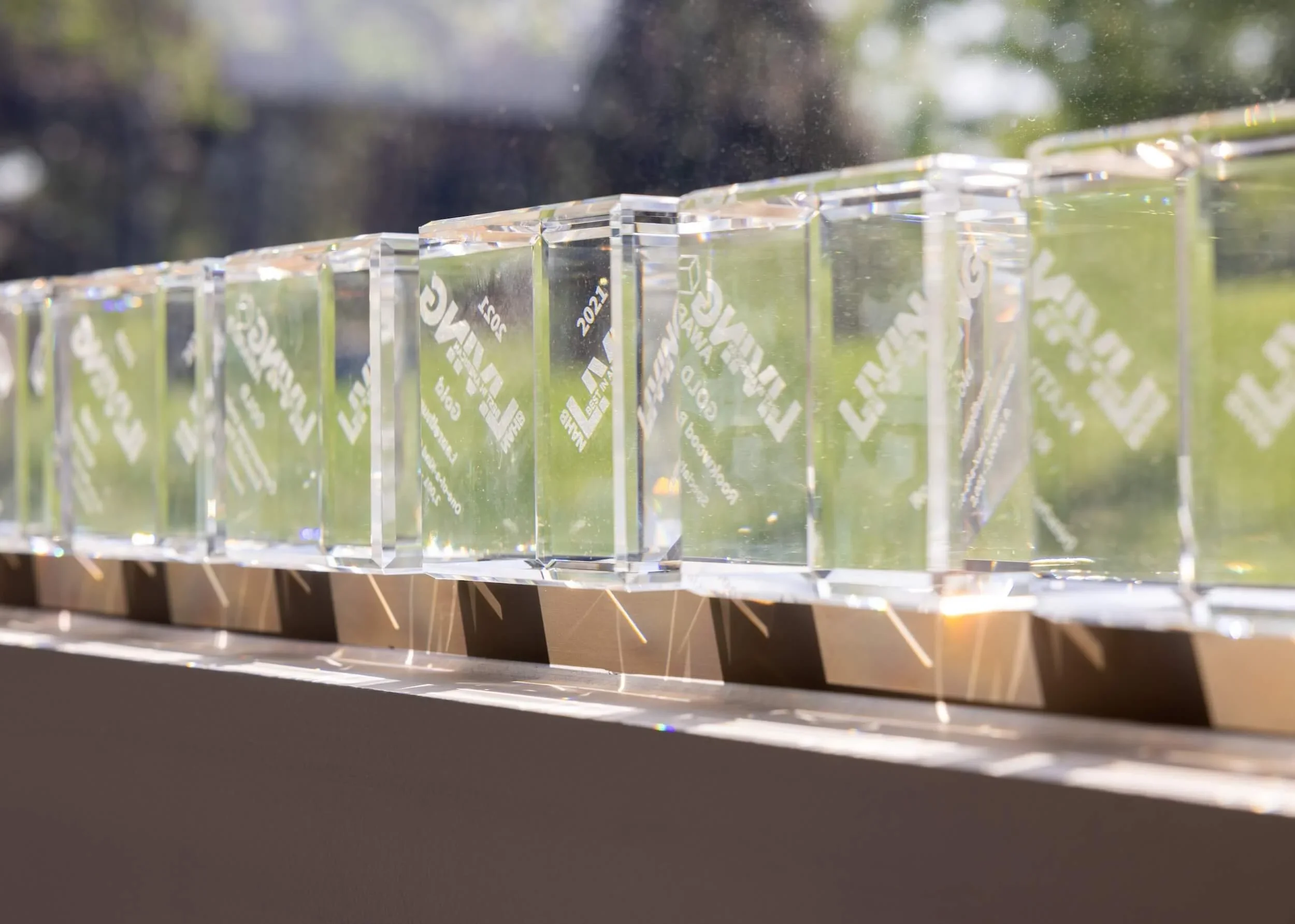 Close-up view of a clear plastic award or trophy with engraved text and logos, resting on a dark surface outdoors, with sunlight creating reflections and a blurred background of trees and sky.
