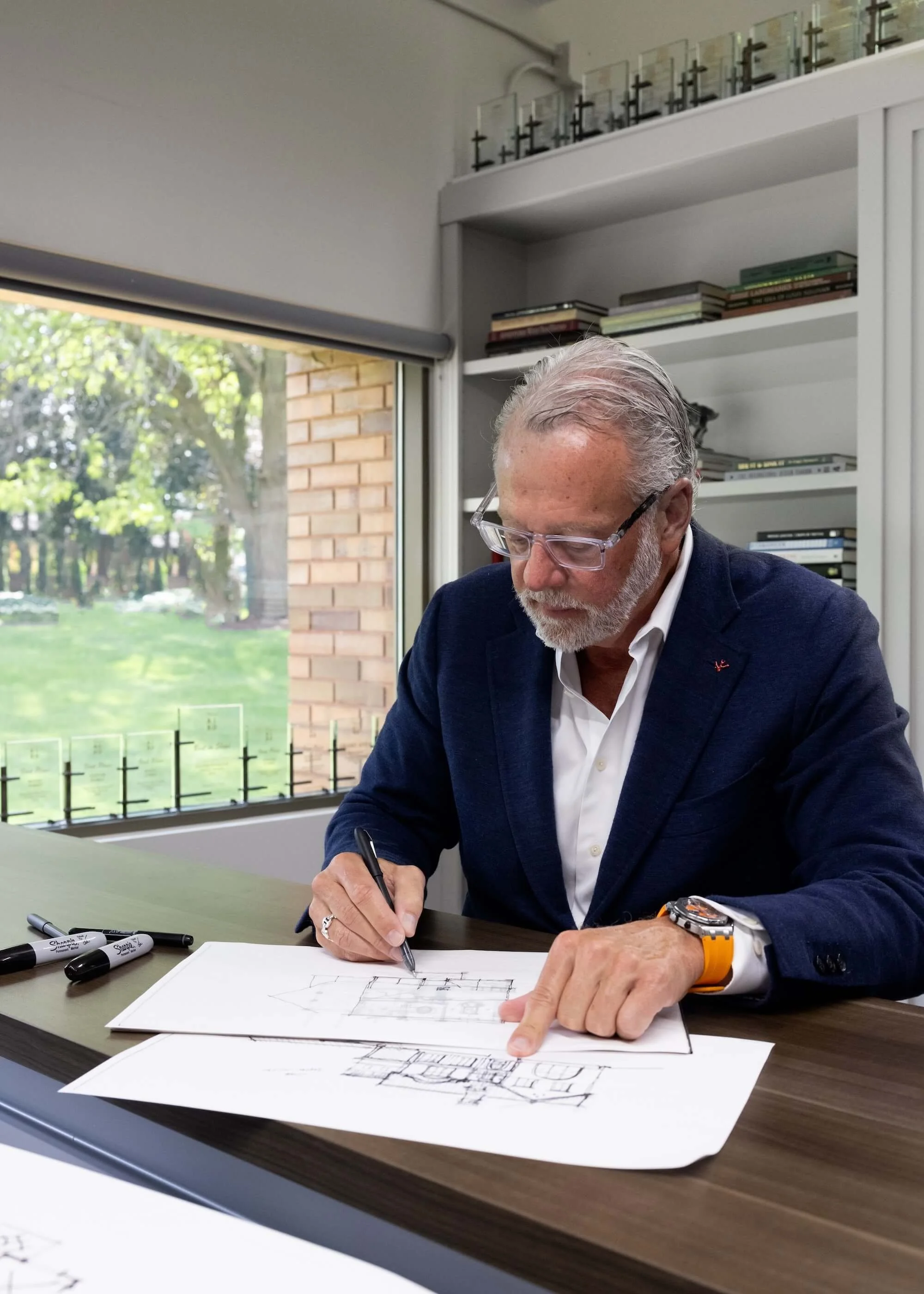 An older man with graying hair and glasses is sitting at a conference table, drawing architectural plans on paper with a black marker. He is wearing a dark blue blazer, a white shirt, and a yellow wristwatch. The room has a large window showing a green outdoor scene and a bookshelf behind him filled with books.