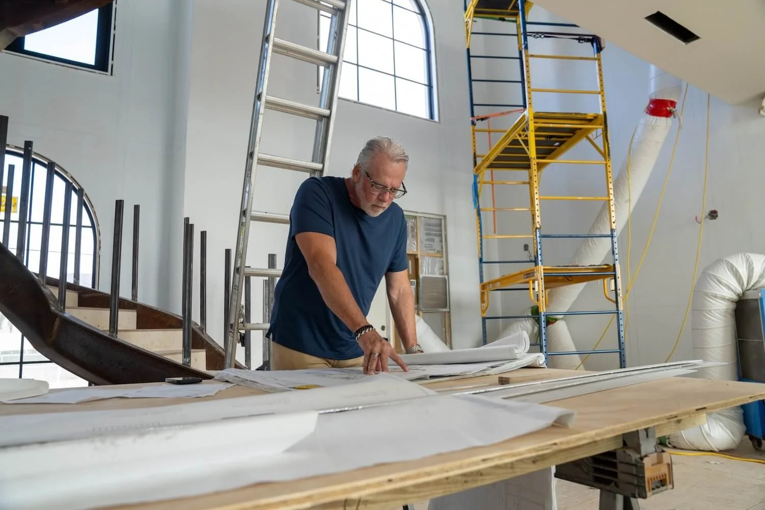 A man with glasses, a beard, and a navy T-shirt is examining blueprints on a table inside a construction site. There are large windows, scaffolding, and construction materials in the background.
