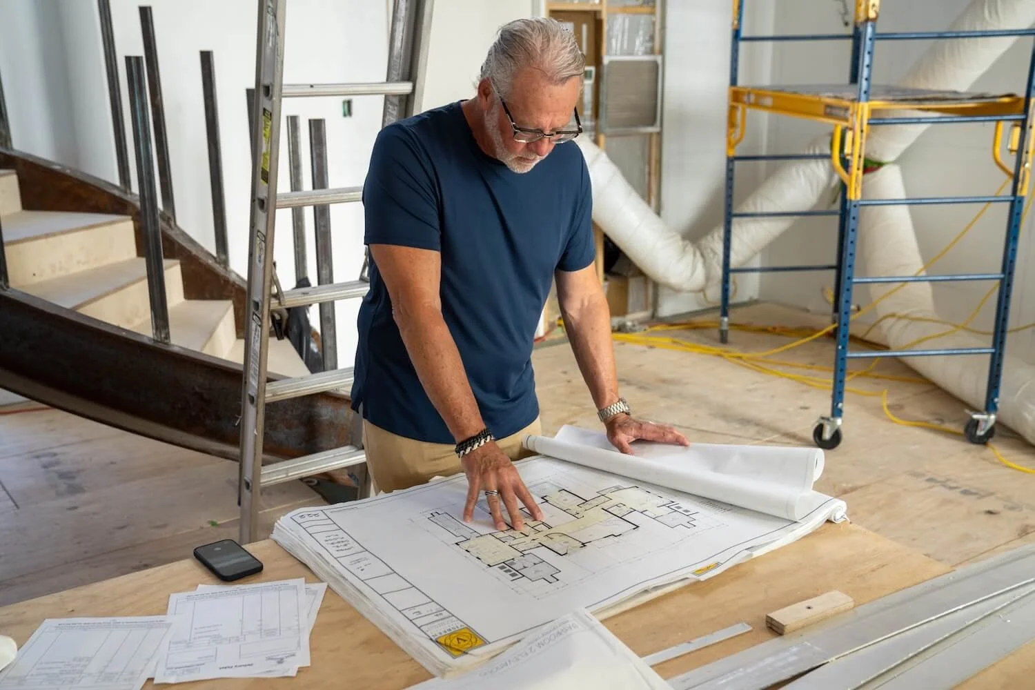 A man in a blue shirt and glasses looking at architectural blueprints on a large wooden table inside a construction site, with a ladder and construction tools and equipment in the background.