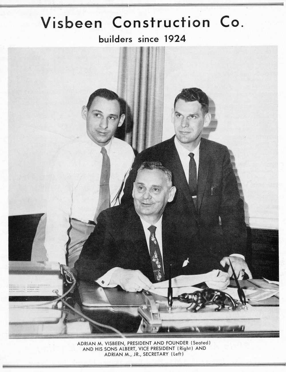 Black and white photo of three men in a conference room, with the caption: Visbeen Construction Co., builders since 1924. Seated is Adrian M. Visbeen, president and founder, with his sons Albert, vice president, and Adrian M. Jr., secretary. The men are dressed in formal business attire, with the seated man wearing a suit and tie. There are papers, pens, and office equipment on the desk, and curtains in the background.