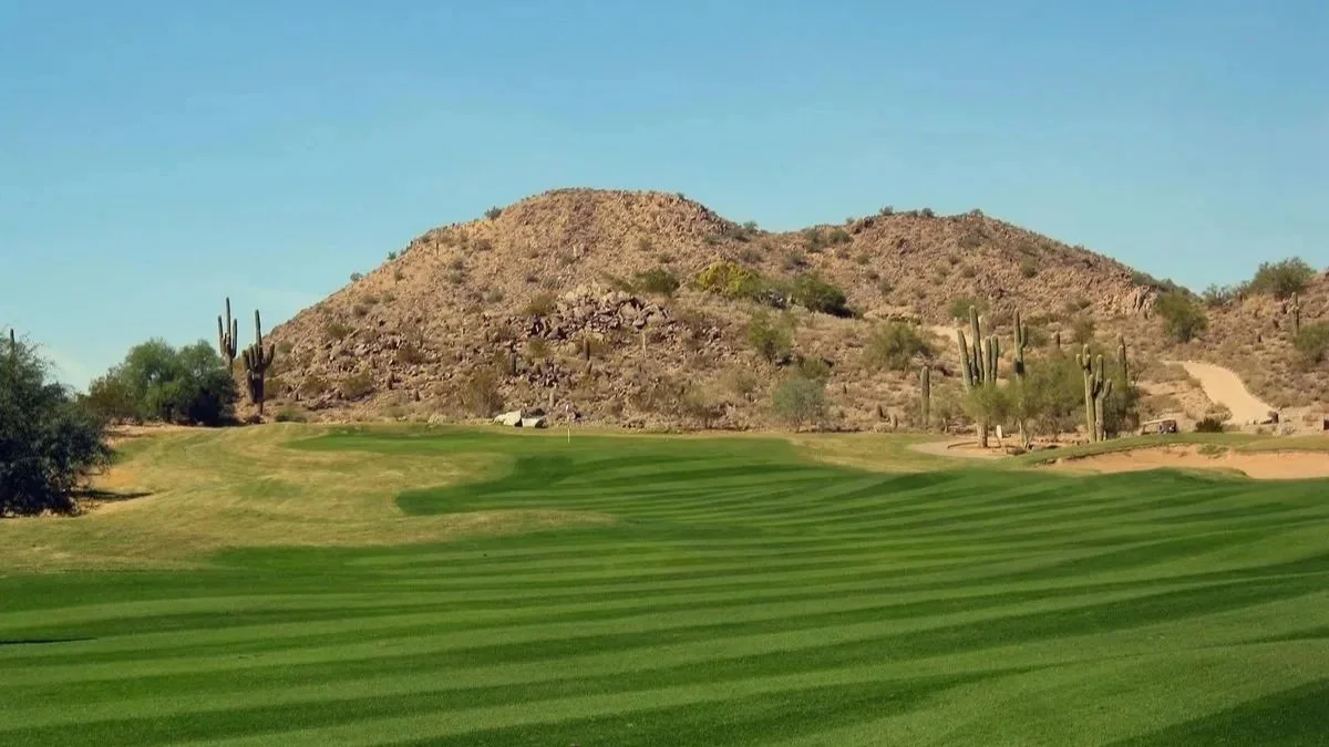 A golf course with well-manicured grass, desert mountains, and cacti in the background under a clear blue sky.