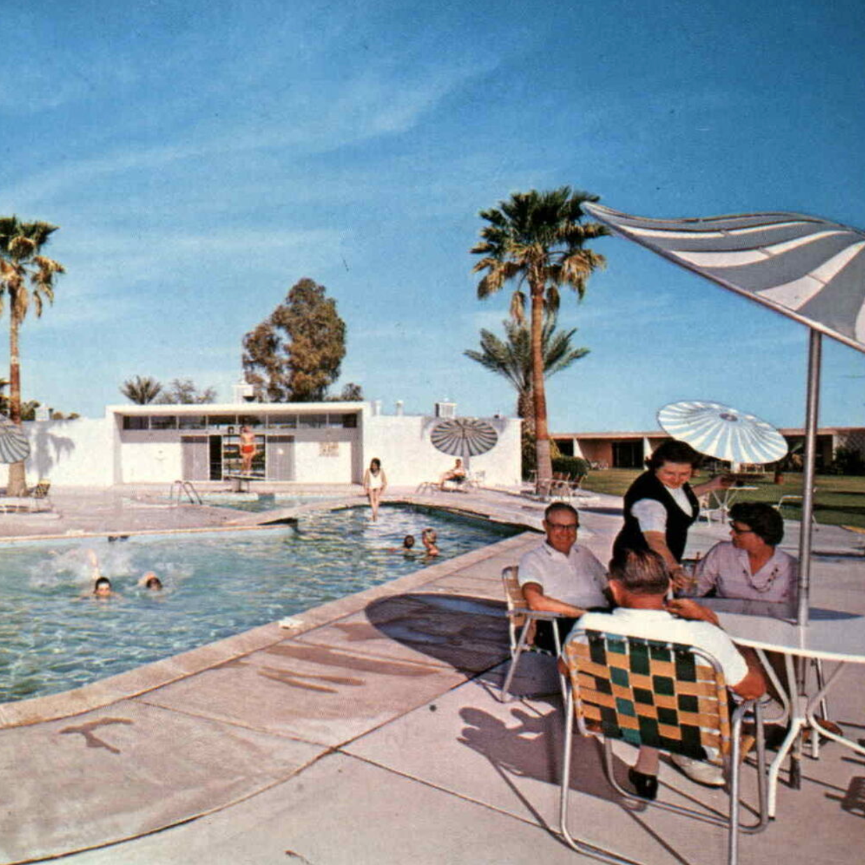A vintage color photograph of a mid-20th century outdoor swimming pool scene with people swimming and relaxing, palm trees, and people sitting at a table under umbrellas.