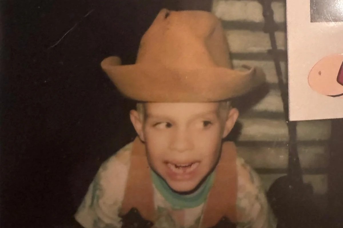 A young boy wearing a cowboy hat and a camouflage shirt, smiling happily, with a brick wall and a poster with a cartoon pig in the background.