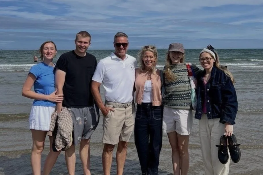 Six people standing on the beach by the ocean, smiling for the camera with waves in the background.