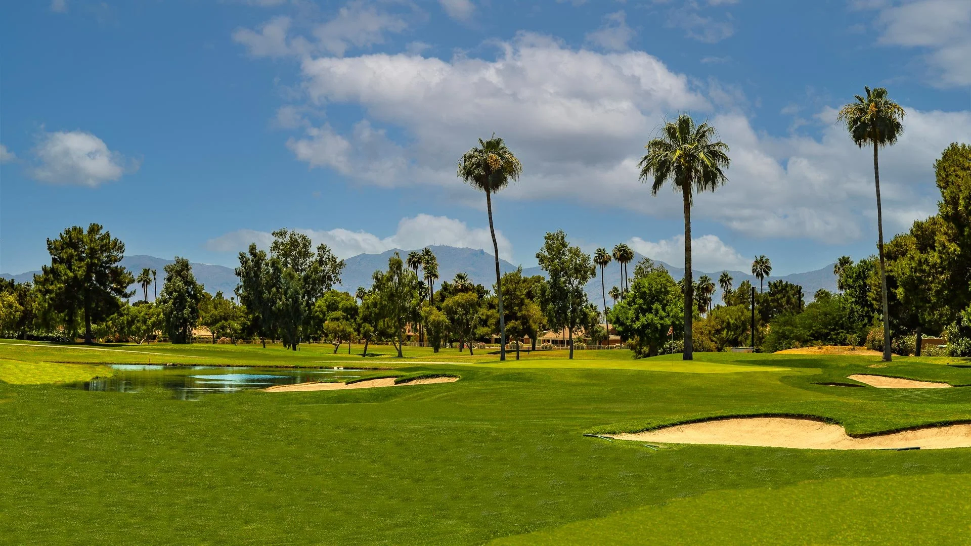 A golf course with lush green grass, sand bunkers, a water hazard, and tall palm trees under a partly cloudy blue sky with distant mountains.