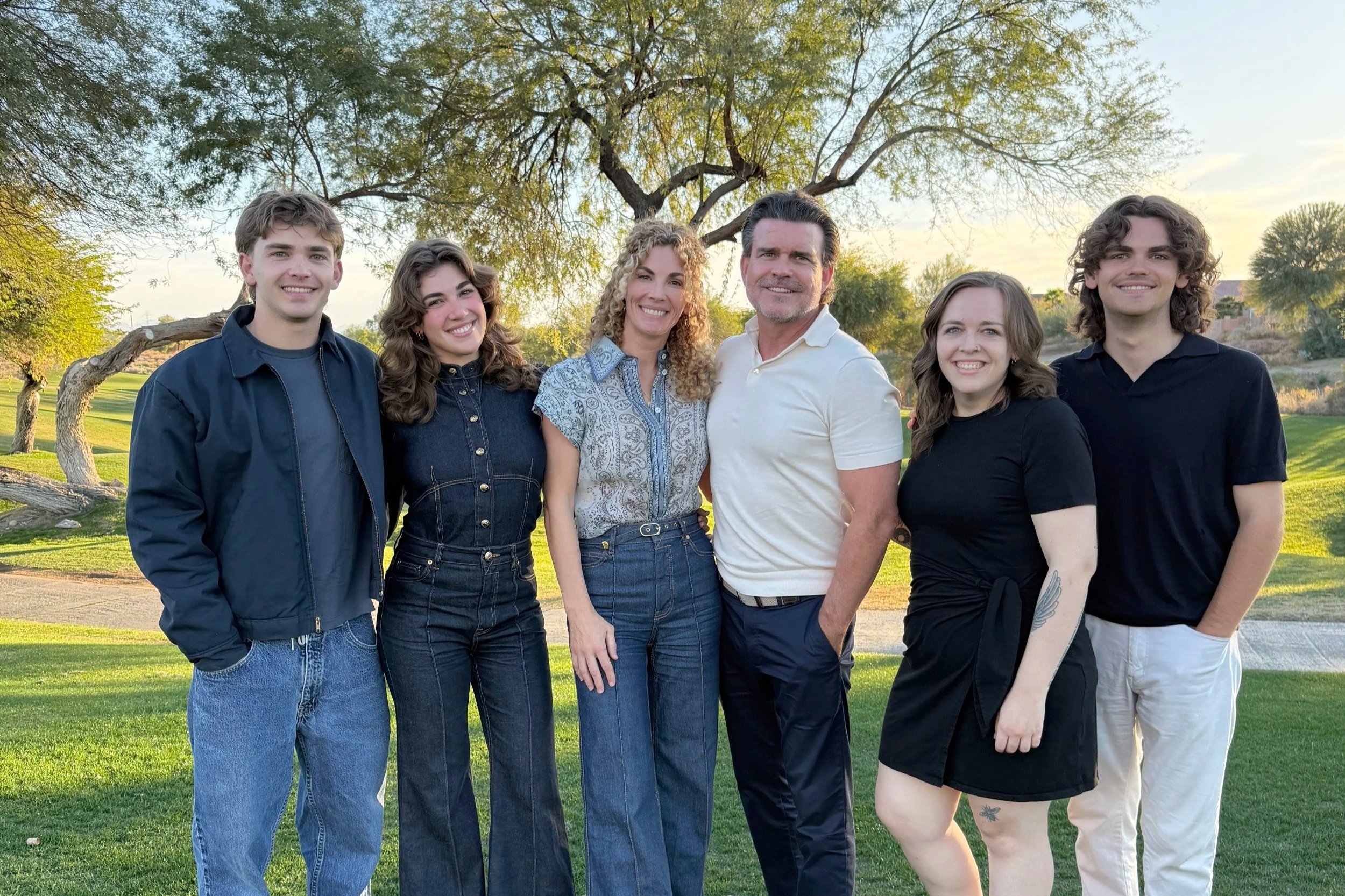 A family of six people standing together outdoors on a sunny day, with trees and grass in the background.