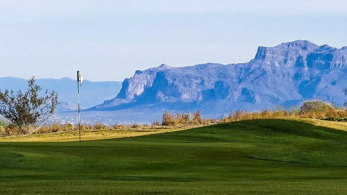 A golf course with green grass and a flagstick on the green, with mountains in the background under a clear sky.