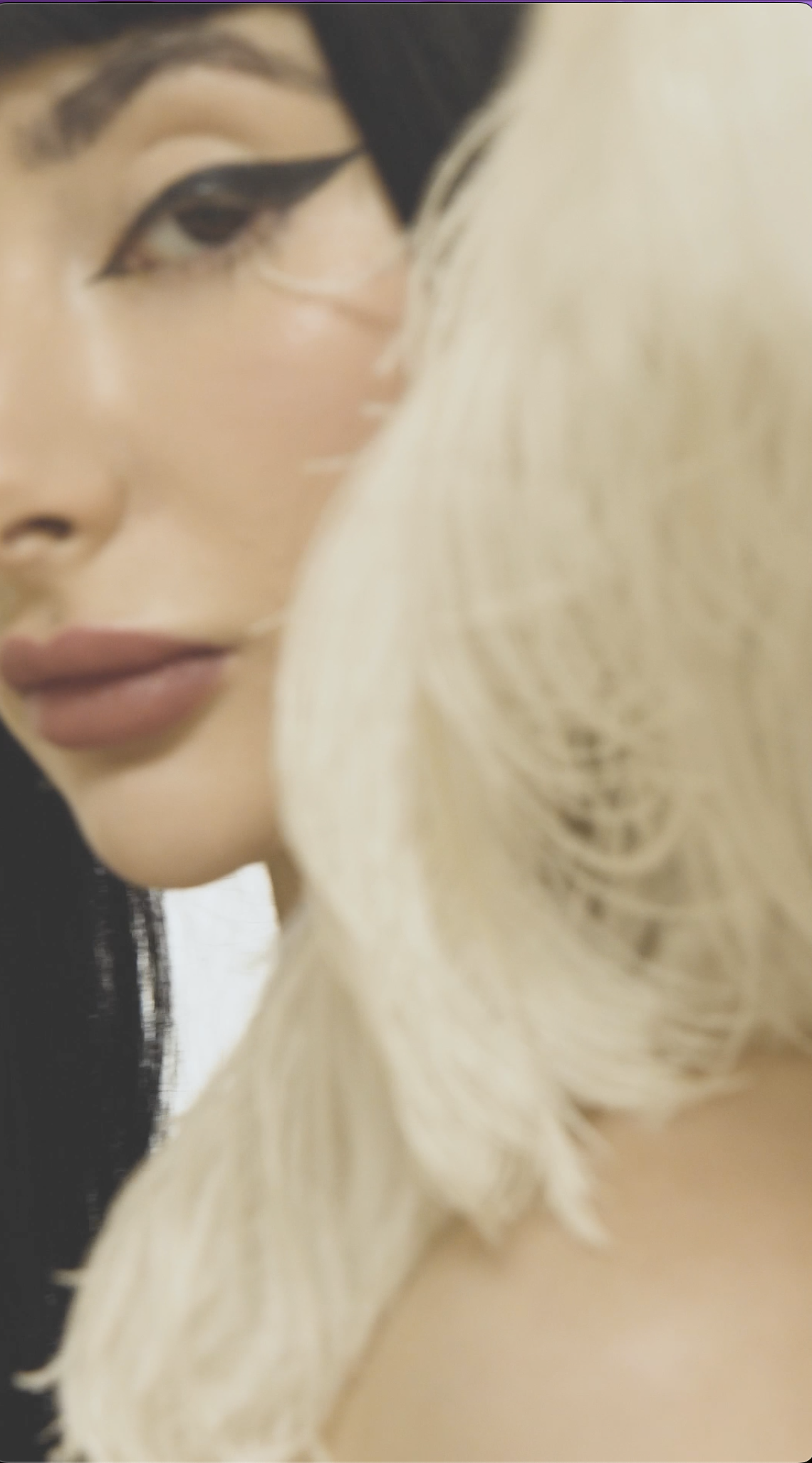 Close-up of a woman with makeup and black eyeliner, complemented by long platinum blonde hair partially covering her face.