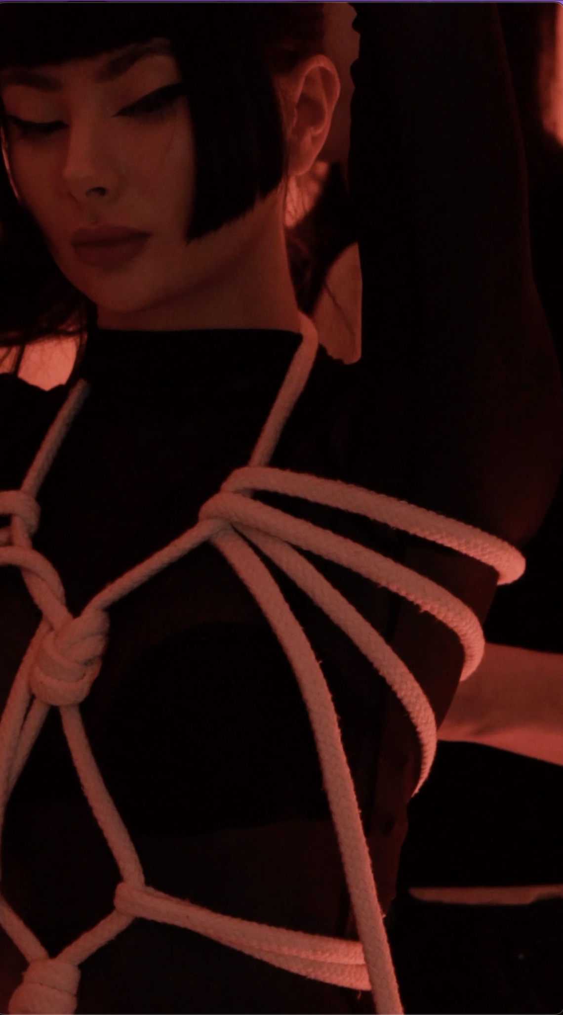 Woman with dark hair styled in a short bob, closed eyes, and makeup, wearing a black top and a hoodie, with white ropes tied around her body in a decorative pattern, in a dimly lit red ambiance.