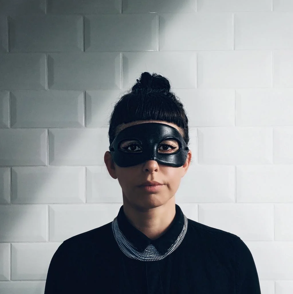 A young woman with dark hair in a bun wearing a black mask and a black collared shirt with silver beaded details, standing in front of a white tiled wall.