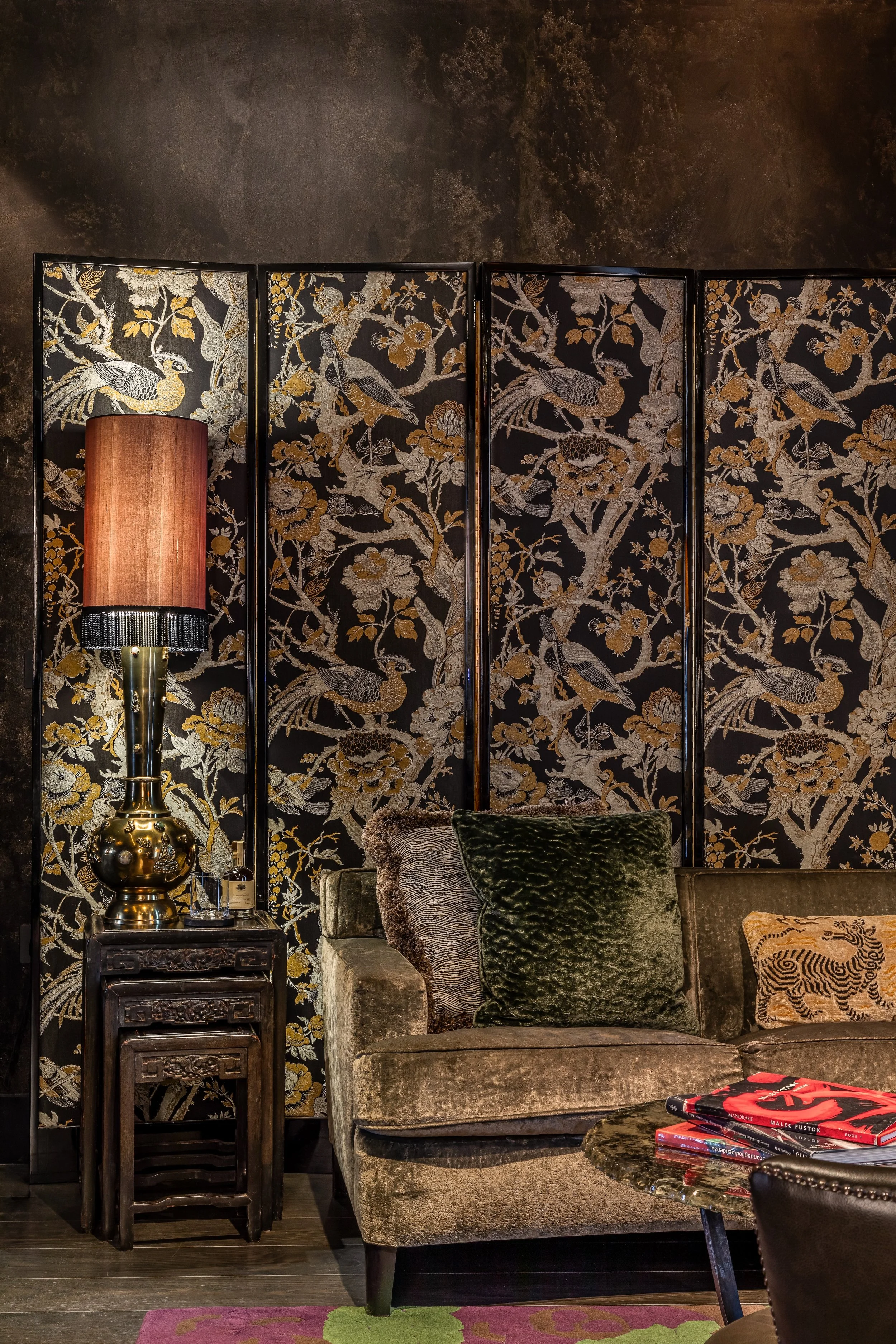 Living room with ornate folding screen decorated with bird and floral patterns, a green velvet pillow on a vintage velvet sofa, a table with stacked jewelry and books, and a large lamp with a rose-colored shade.