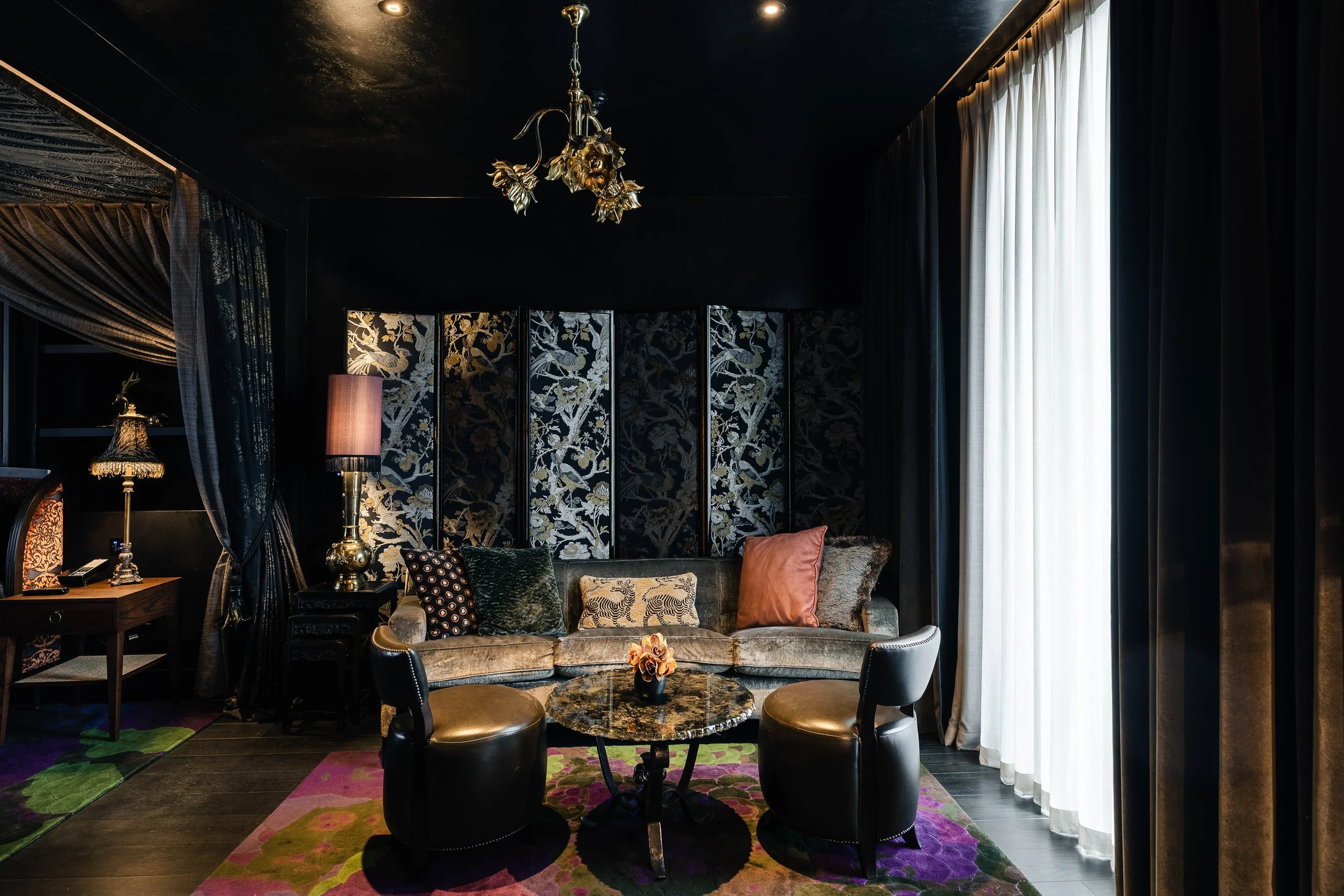 Luxurious living room with dark walls, ornate chandelier, velvet sofa with decorative pillows, small round marble table, two black leather chairs, floral patterned rug, large window with white curtains, additional lamps and curtains on the sides.