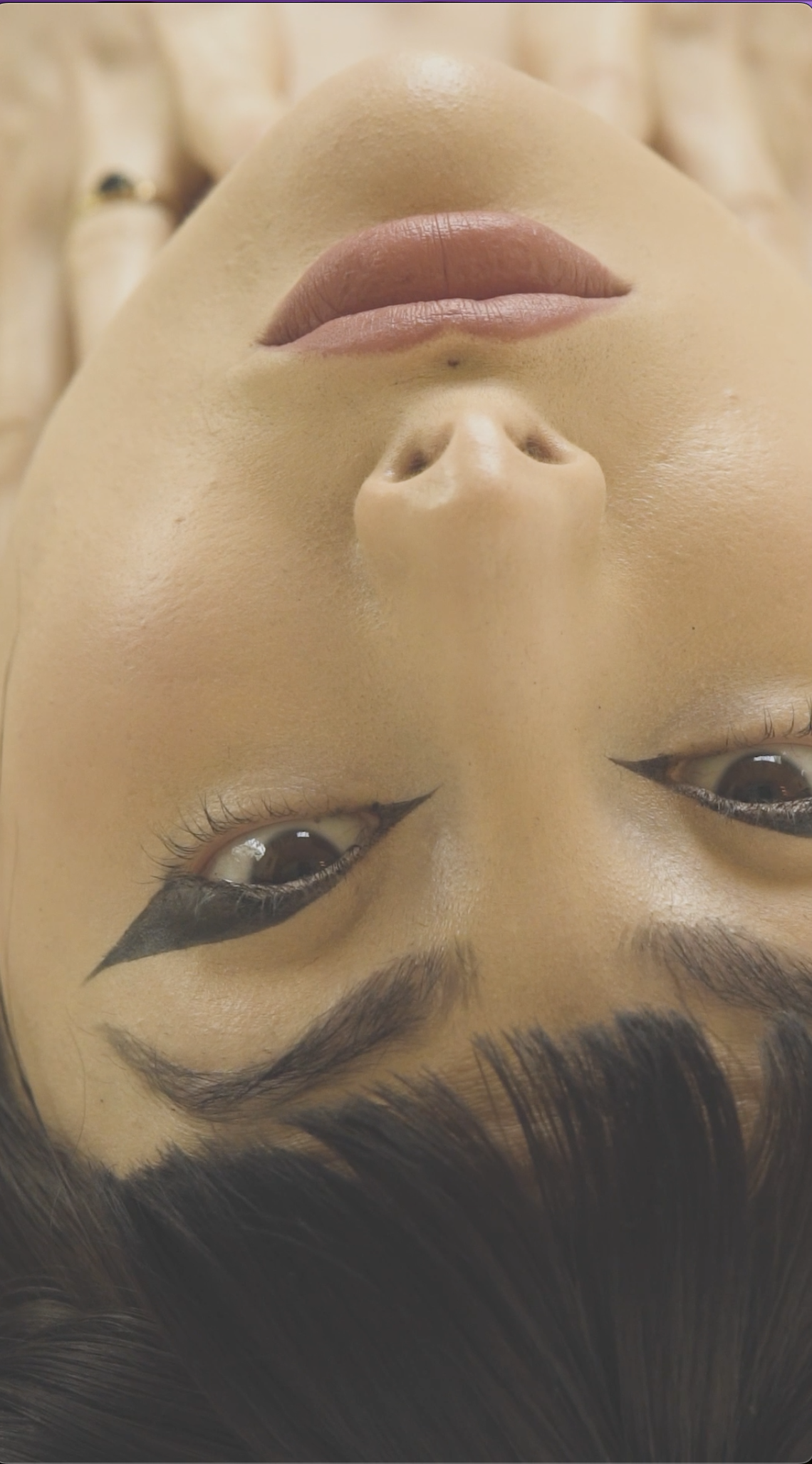 Close-up of a person's face lying upside down with makeup, including winged eyeliner and glossy lips.