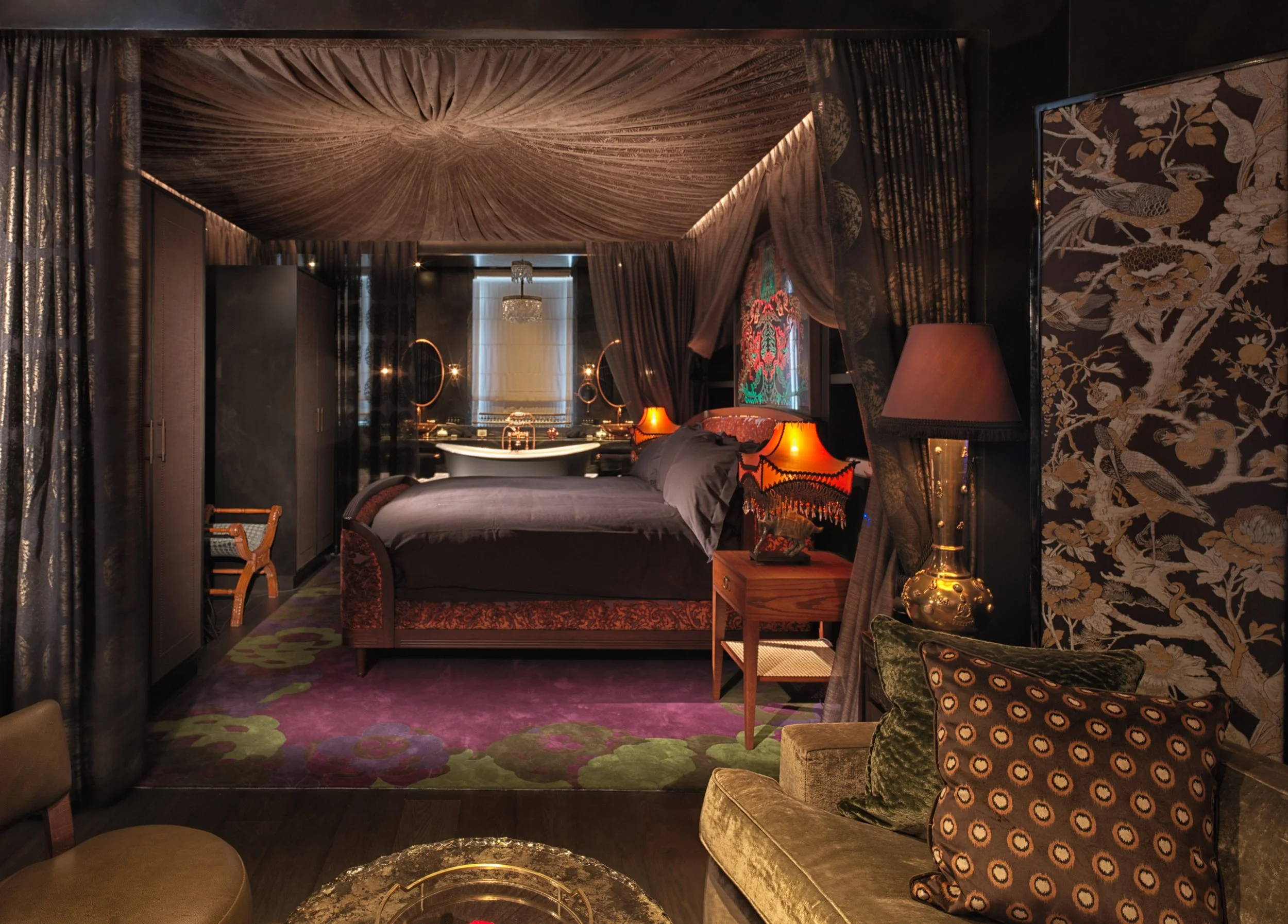 A luxurious bedroom with dark decor, purple curtains, and ornate furnishings. The bed has dark bedding and there are vintage lamps on side tables. The room features patterned wallpaper and a plush green sofa with decorative pillows.