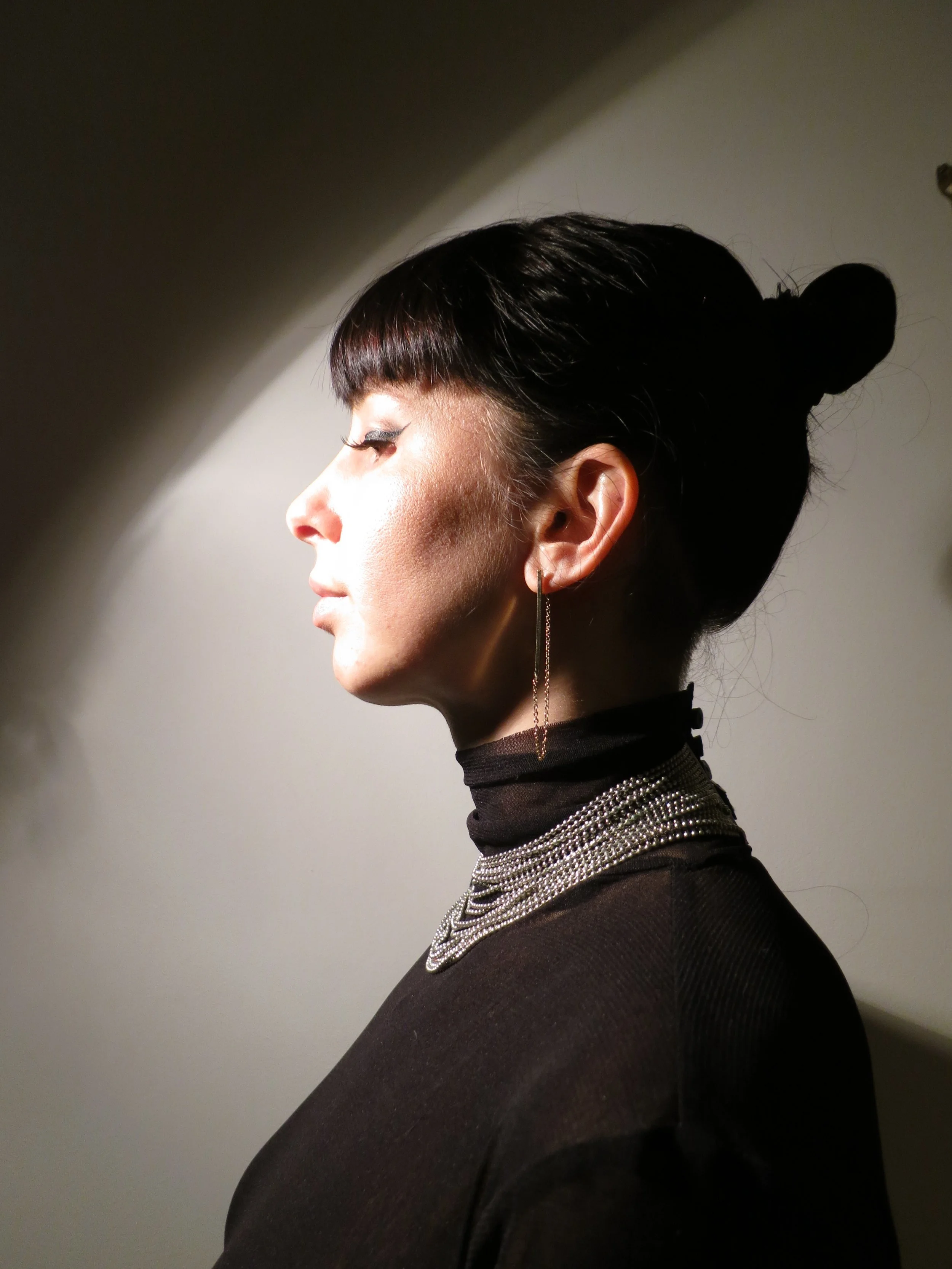 Side profile of a woman with dark hair styled in an updo, wearing a black turtleneck, layered necklaces, and a single long earring, with lighting creating shadows on the background.