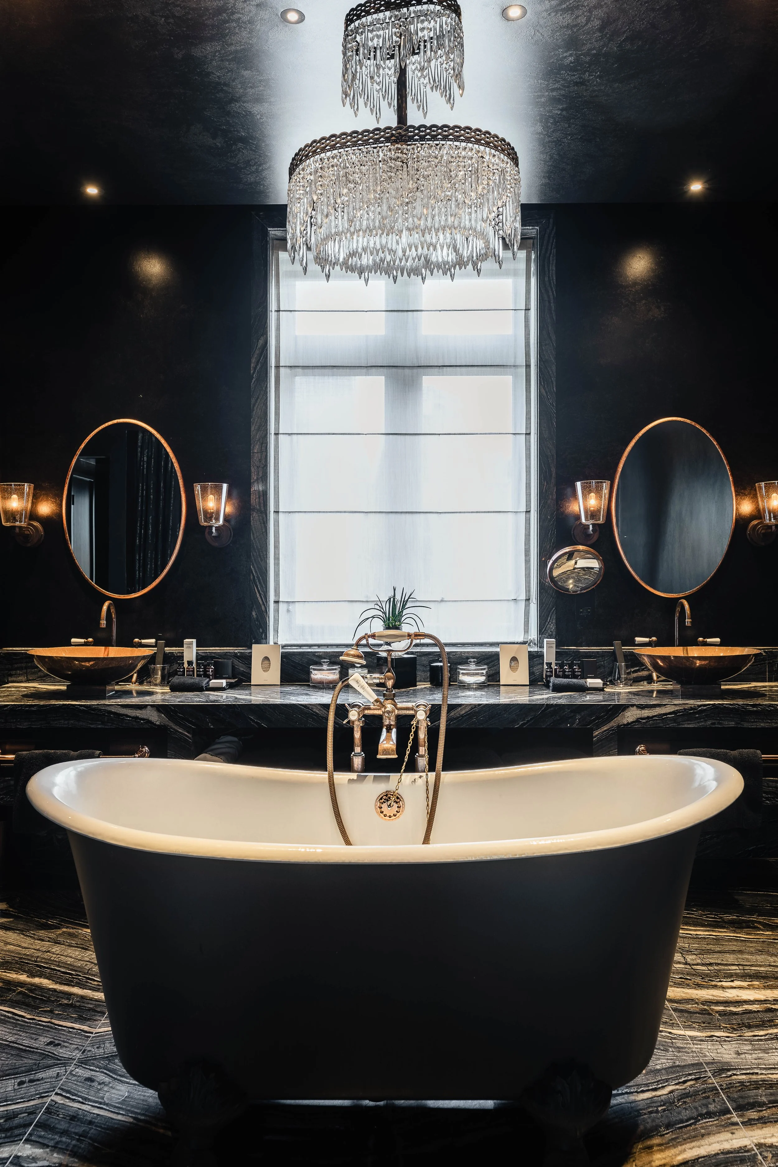 Luxurious bathroom featuring a classic clawfoot bathtub in the foreground, a black marble vanity with two sinks and oval mirrors, a large window with sheer curtains, and a grand crystal chandelier hanging from the ceiling.