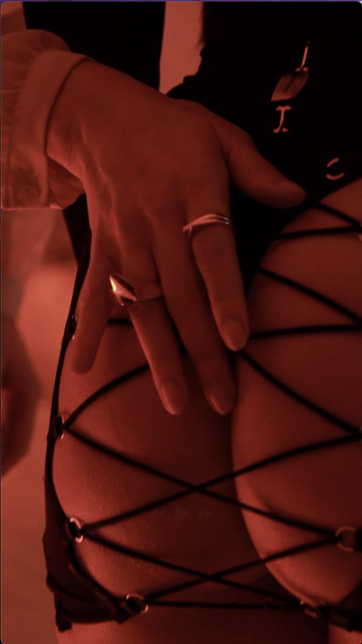 A person wearing black lingerie with a crisscross design, showing a hand with rings and a watch, in a dimly lit, red-tinted environment.