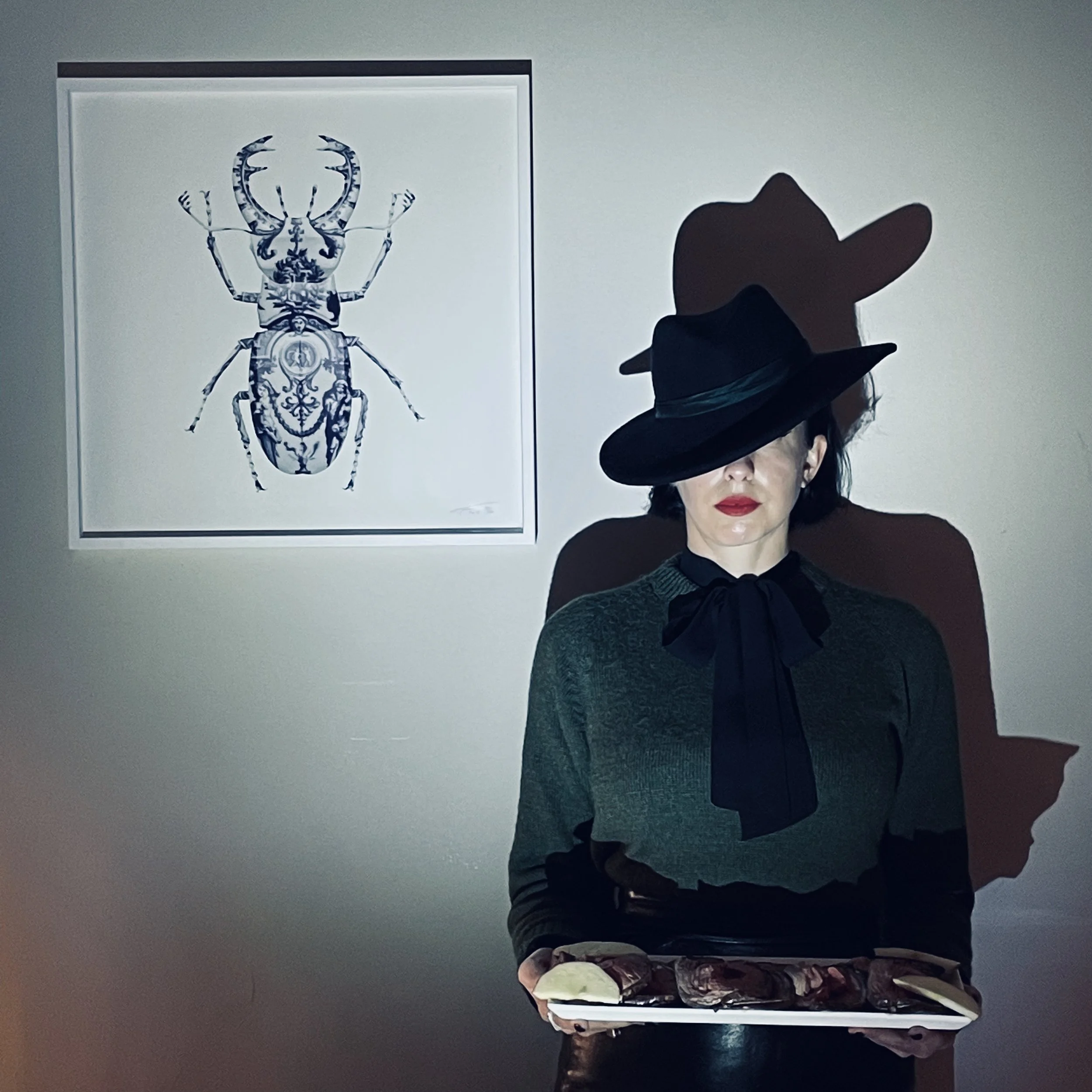 A woman with red lipstick wearing a large black hat, a dark green top, and a black bow tie, holding a tray with food, standing in front of a wall with a framed artwork of a beetle.