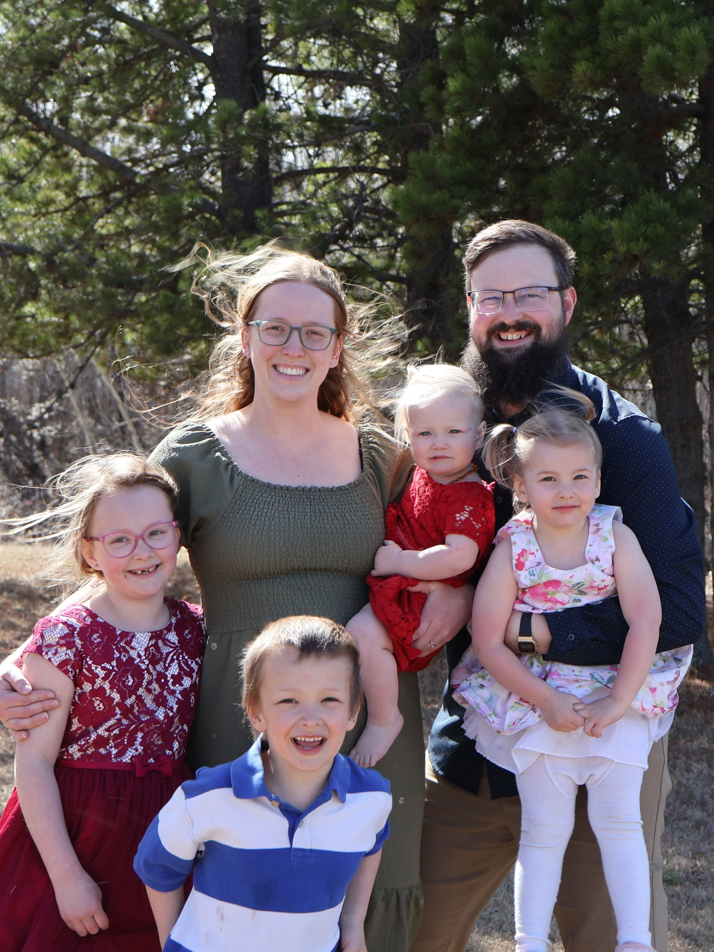 A family portrait of adults and children outdoors with trees in the background, smiling at the camera.