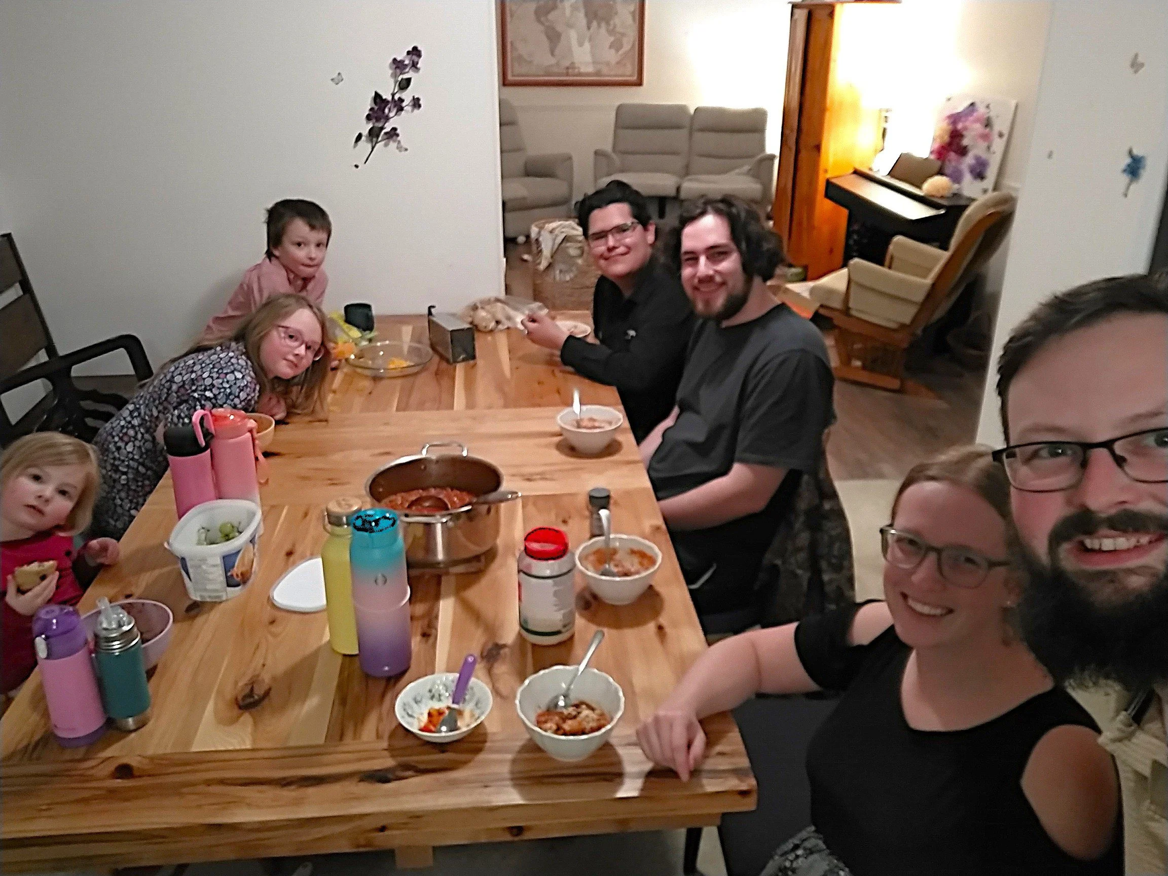 Family gathered around a wooden dining table enjoying a meal in a cozy home, with a background of couches and a piano.