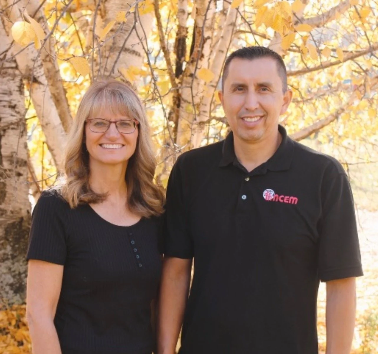 A woman and a man standing outdoors in front of a tree with yellow autumn leaves, smiling at the camera.
