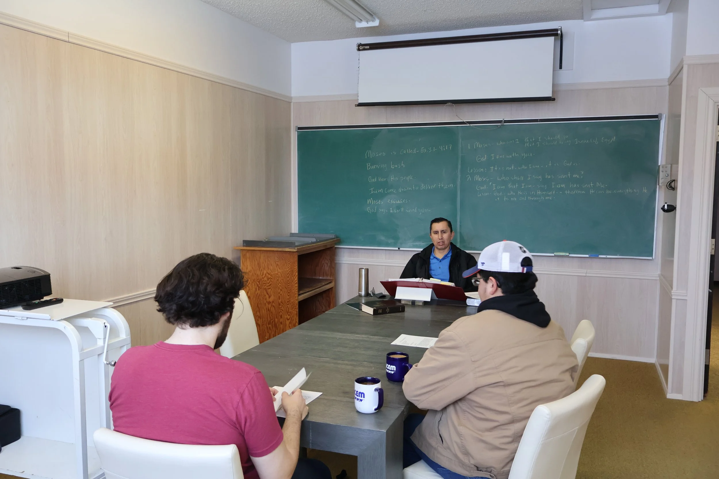 A classroom scene with three men sitting at a table and one instructor at the front. The instructor is sitting at a desk near a chalkboard filled with notes, and the men are reading and taking notes. The room has a white wall, a projector on a cart, and coffee mugs on the table.