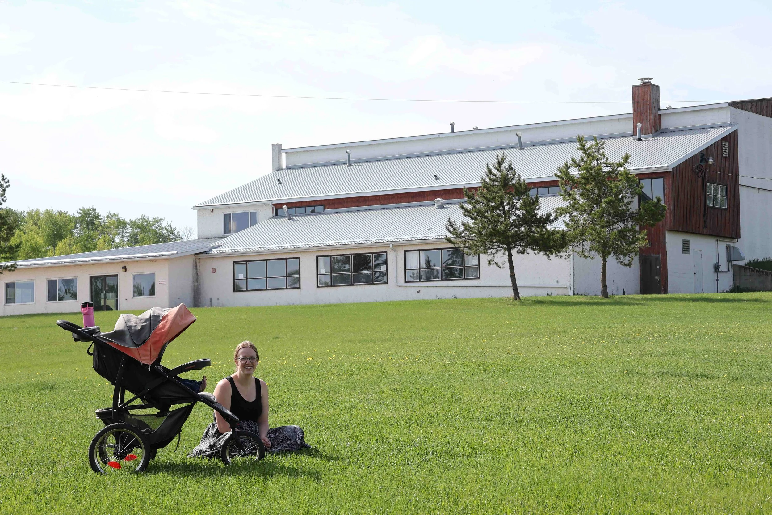 A woman sitting on the grass next to a stroller on a large green lawn, with a modern multi-story building and trees in the background.