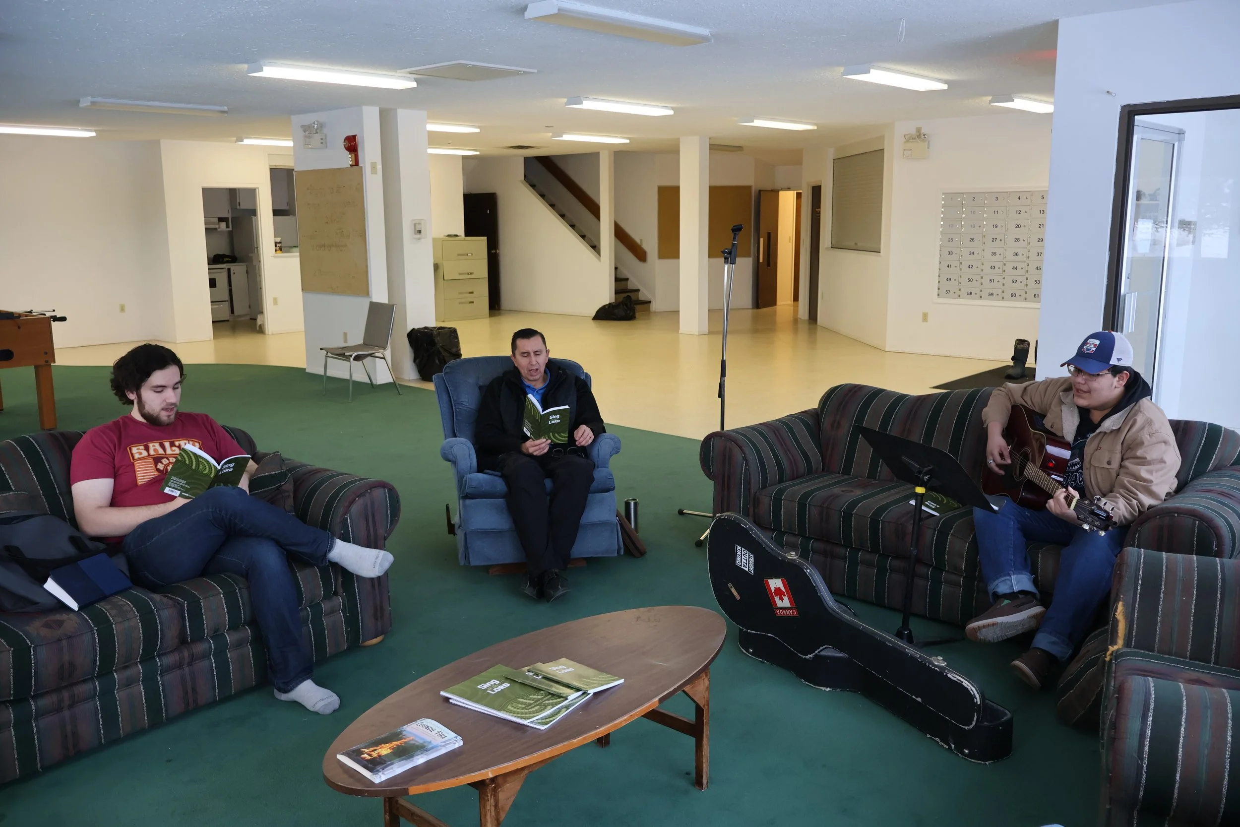 Three men sitting on sofas and a chair in a spacious room. The men are singing and one is playing guitar. A guitar case lies open on the floor, and a coffee table holds a few books.
