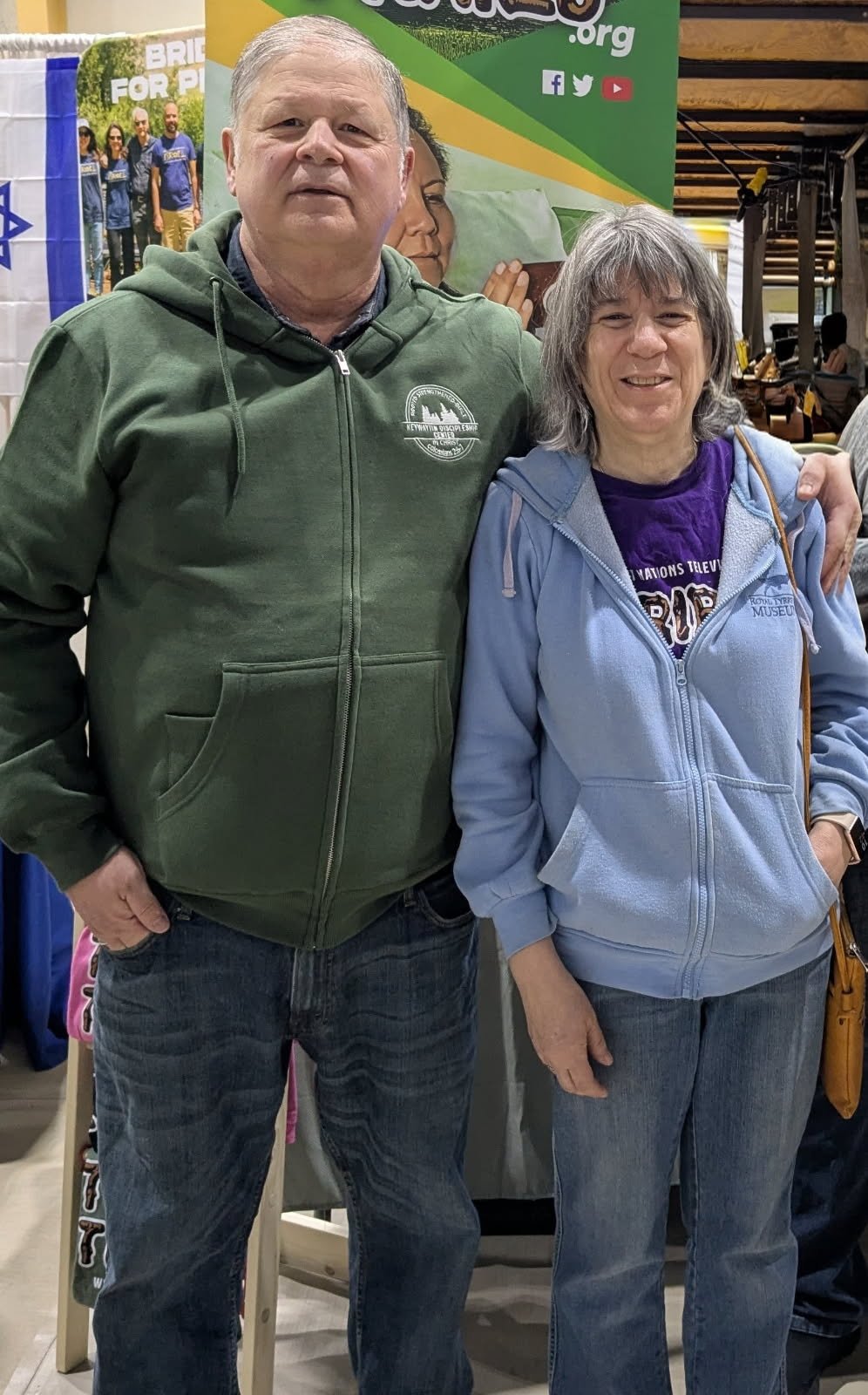 A man and woman standing side by side indoors, smiling at the camera, with a green and yellow promotional backdrop behind them, and other people in the background.