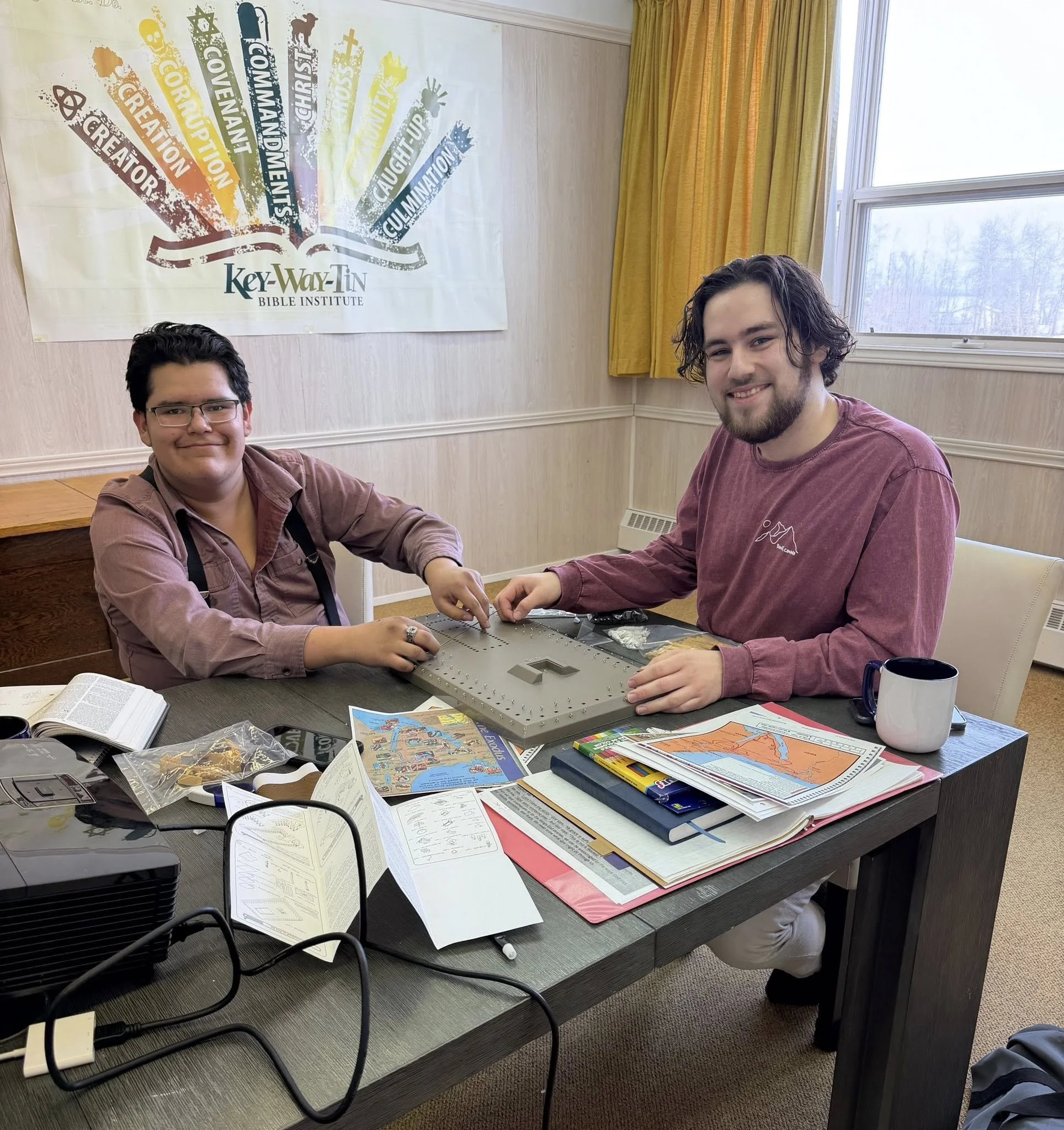 Two young men sitting at a table building a model of the Tabernacle inside a room with a window and a yellow curtain. There are books, papers, and a mug on the table, and a poster on the wall behind them with various colorful words."