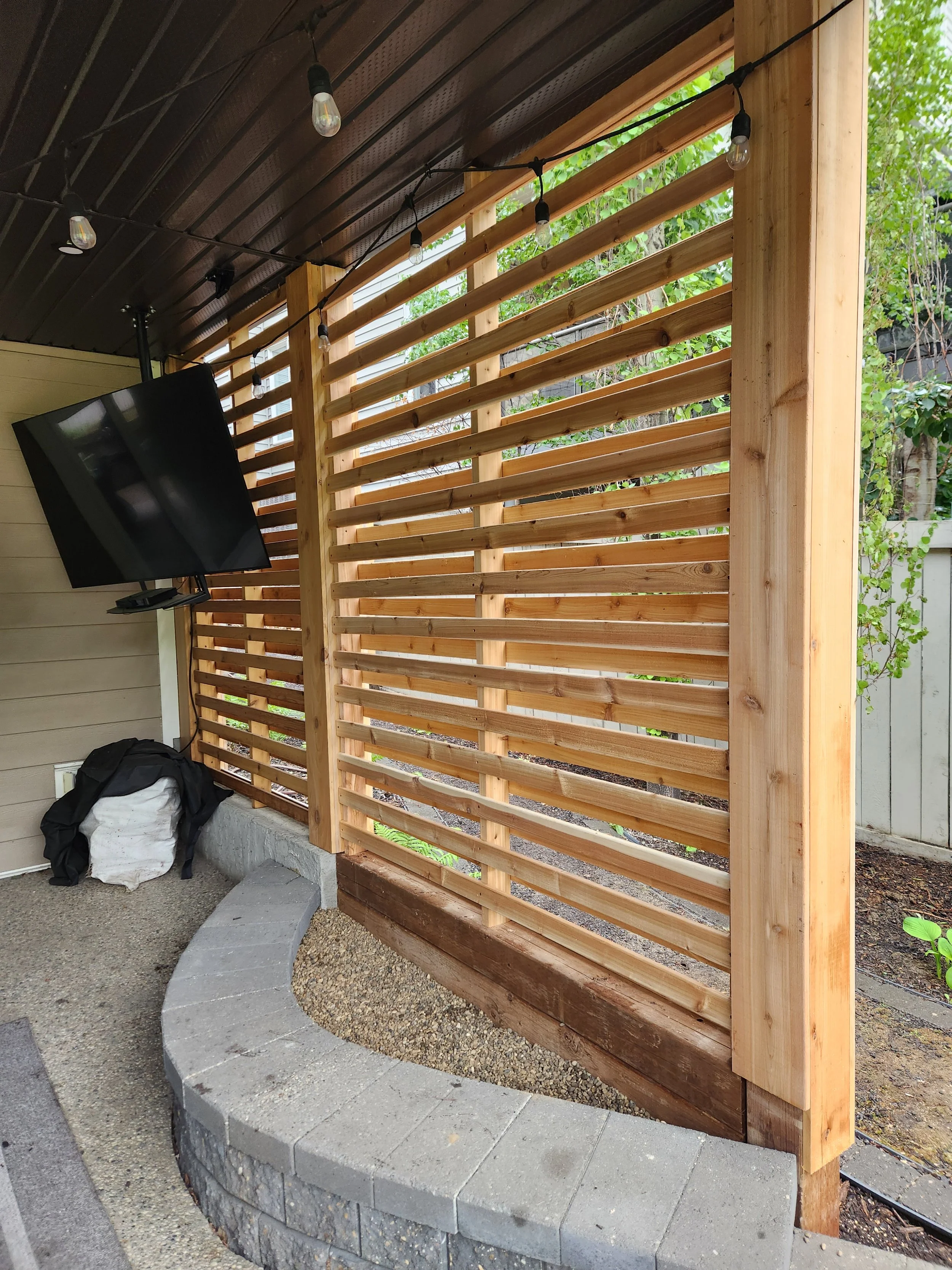 A wooden privacy screen with horizontal slats on a porch, with a mounted flat-screen TV to the left, and a curved stone planter at the base, in an outdoor setting with greenery in the background.