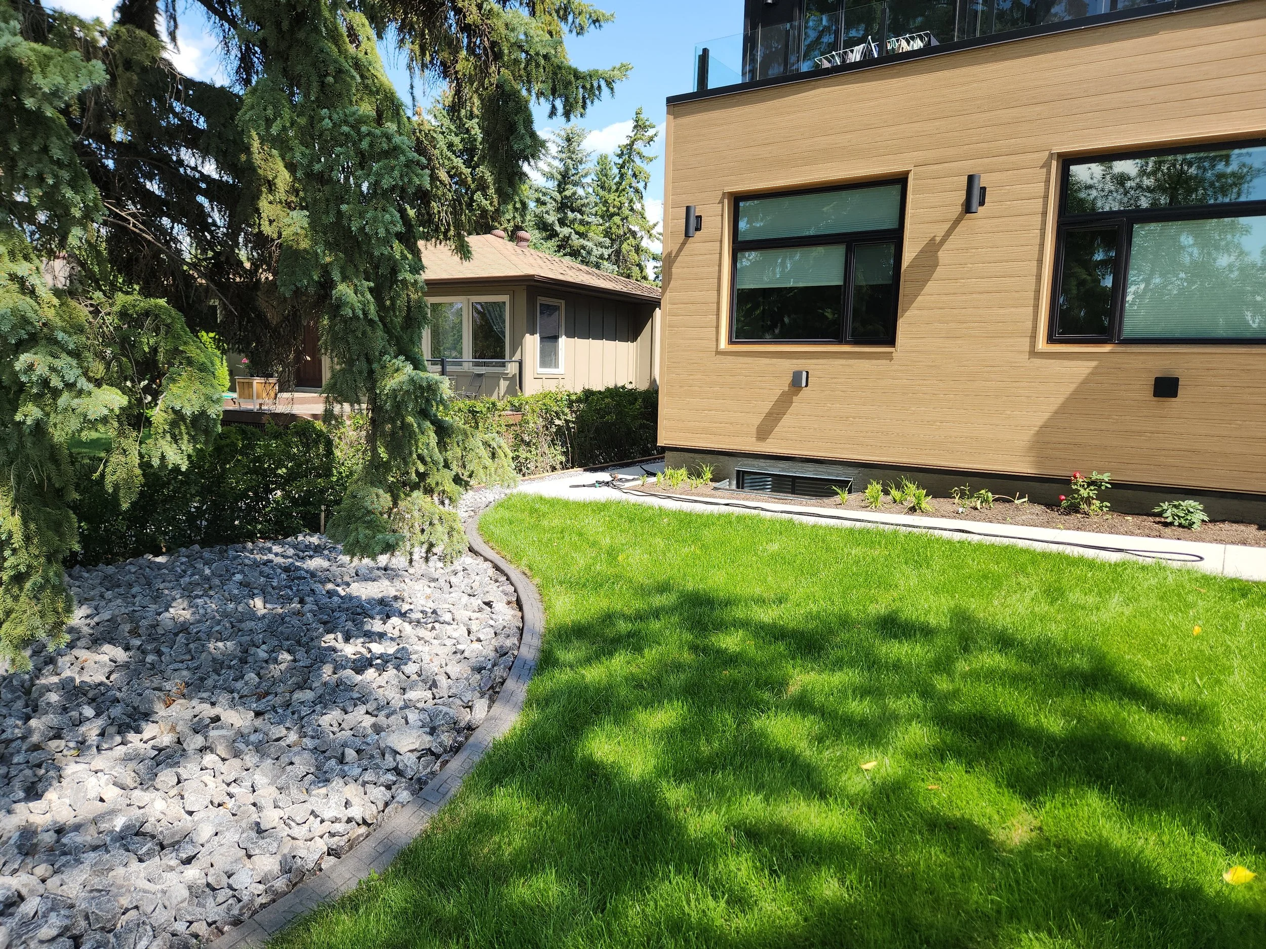 A backyard garden with green grass, a curved bed of rocks, and a modern wooden house with large windows, shaded by trees on a sunny day.